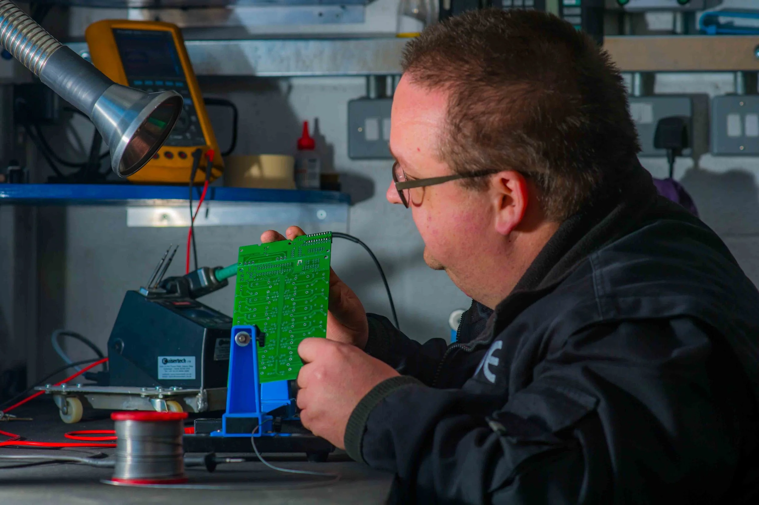 A man working on an electronic circuit board in a workshop setting, with tools and equipment around him.