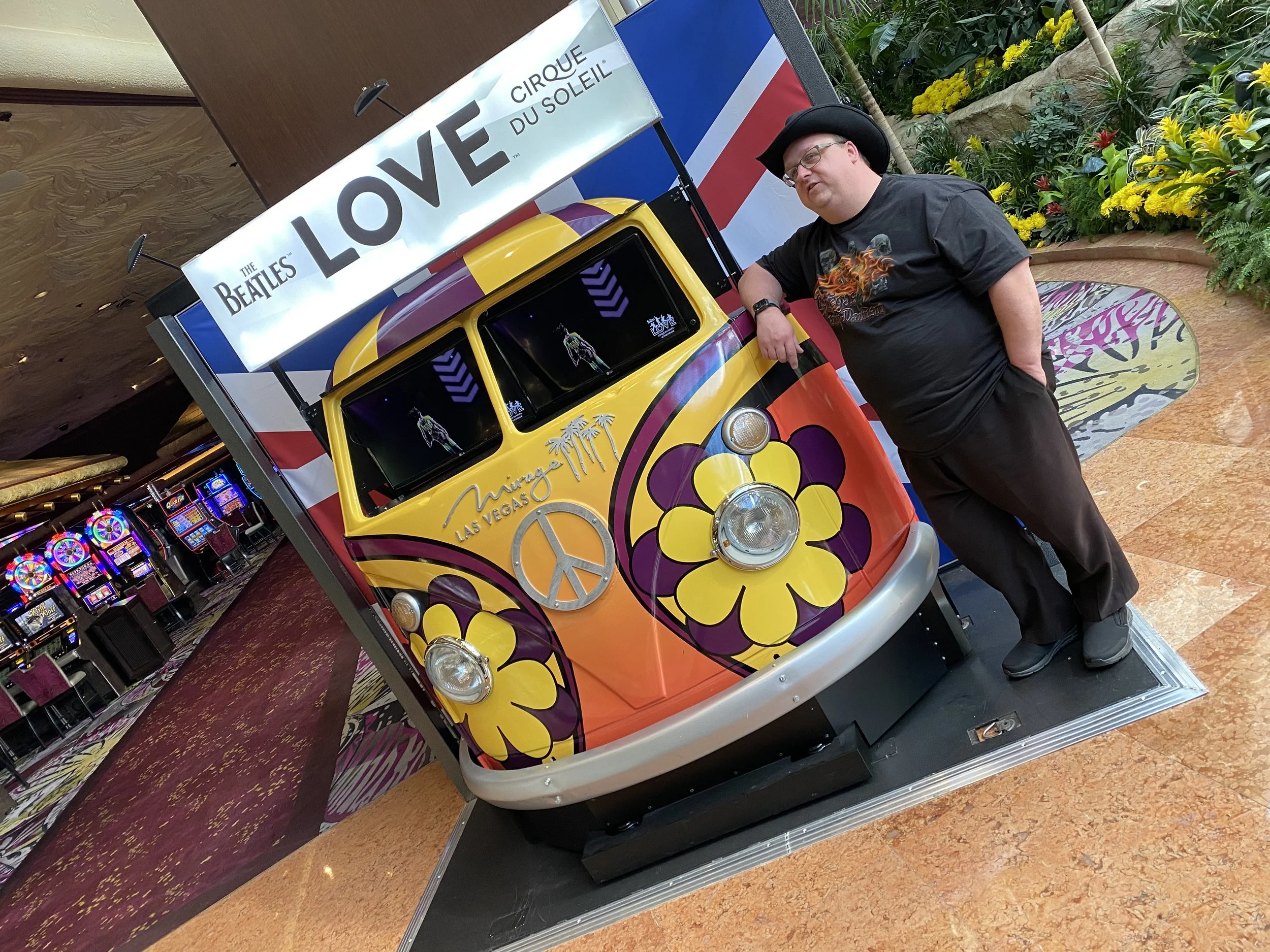 A man wearing glasses, a black cowboy hat, and a black T-shirt with a fiery dragon design, stands next to a colorful vintage Volkswagen van decorated with yellow and purple flowers, and peace symbols, inside a casino or entertainment venue with a lush indoor garden and slot machines in the background.