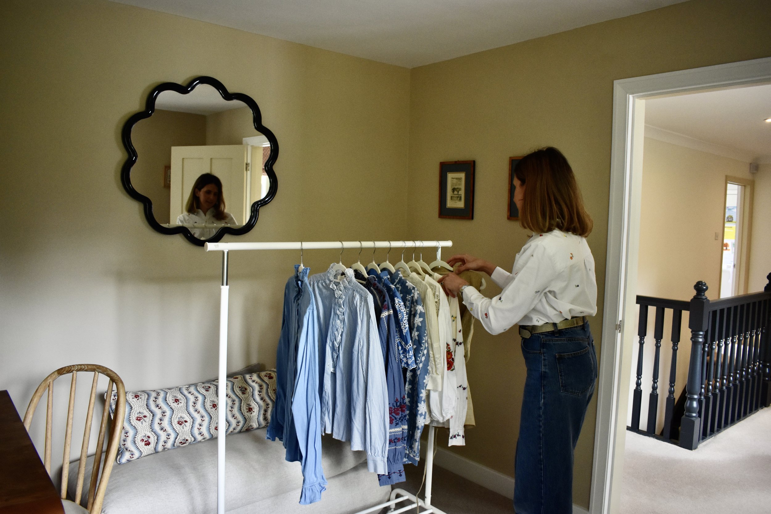 A woman with shoulder-length brown hair, wearing a white embroidered blouse and jeans, stands in a room looking at a clothing rack with various shirts, with her reflection visible in a decorative black-framed mirror on the wall.