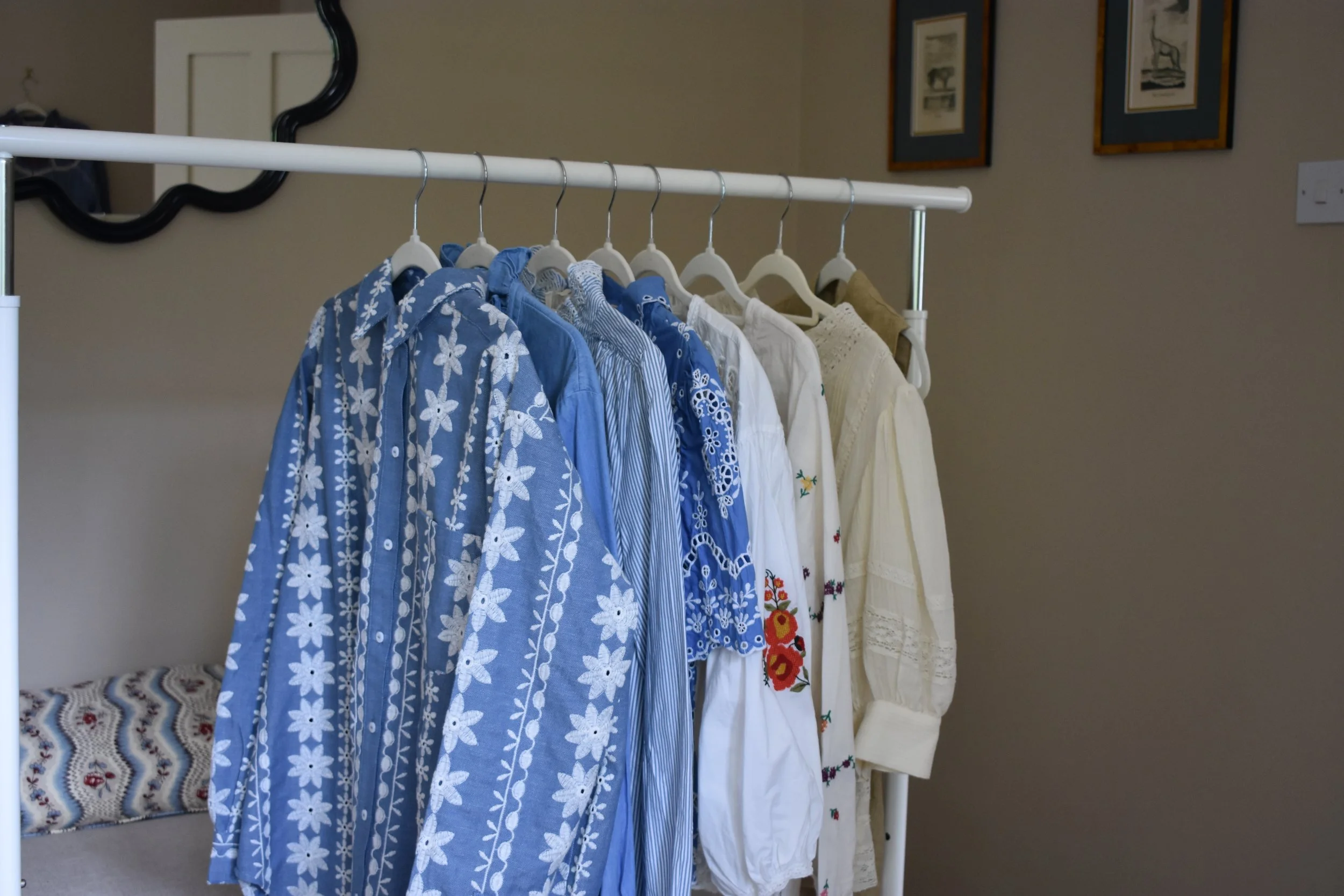 Clothing rack with various blue and white embroidered and patterned tops and dresses, situated in a room with framed artwork on the wall.