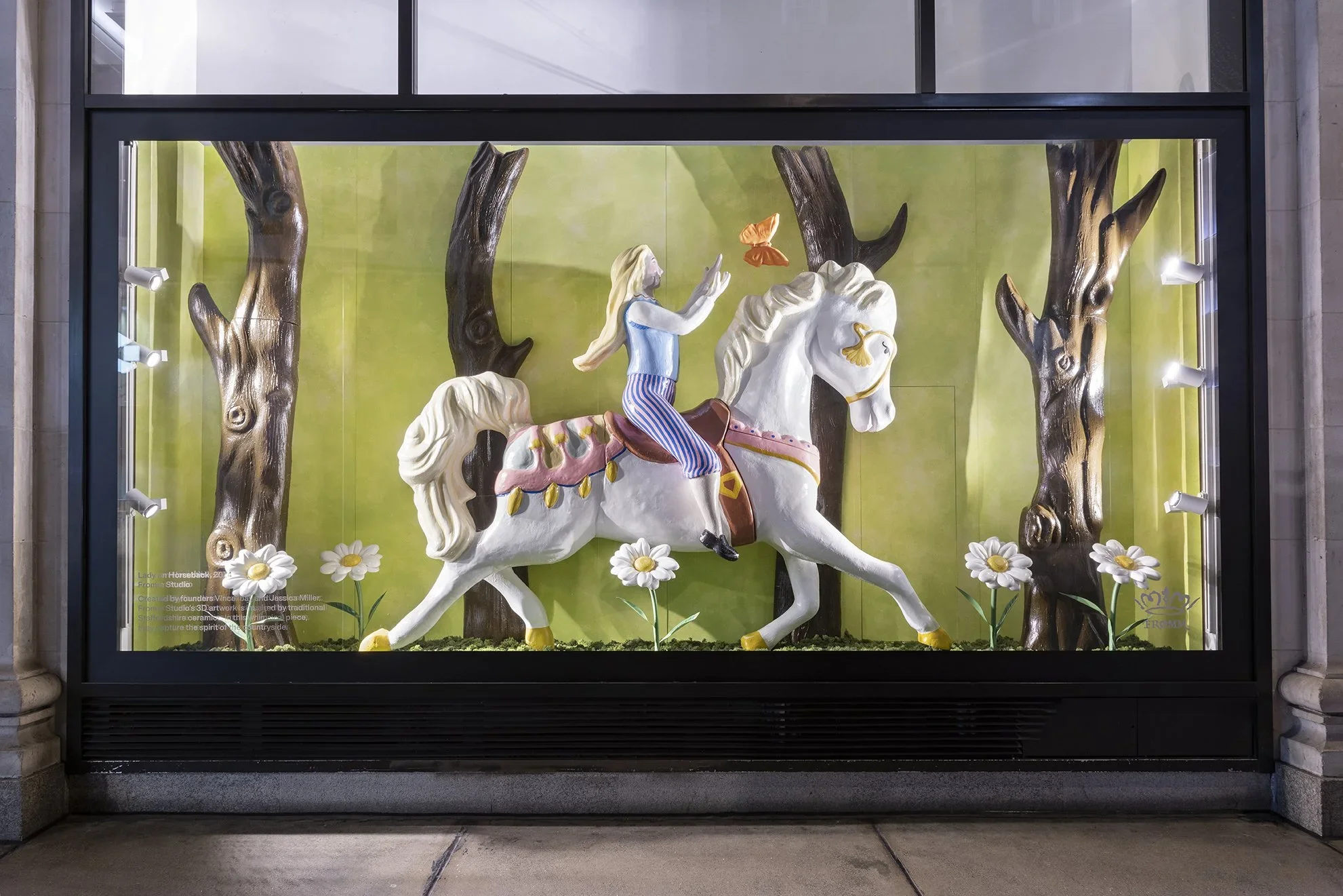 A colorful display featuring a white carousel horse with a blonde woman in striped pants riding it through a forest scene. The scene includes dark brown trees, white daisies, and a green background with a pink butterfly flying nearby.