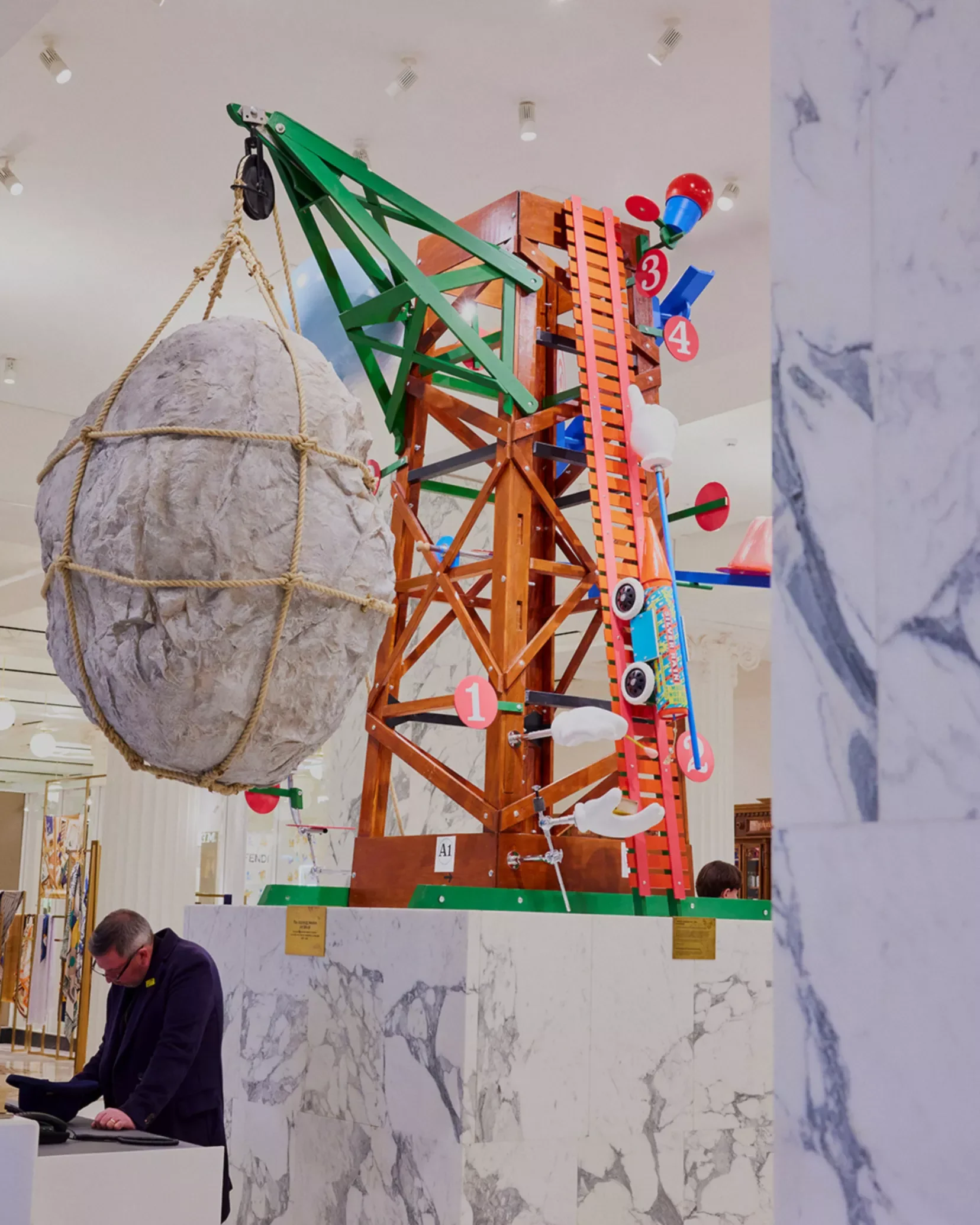 Indoor display with a wooden climbing structure, a hanging large rock held by ropes, and a person in a suit standing at a table, all set against a marble wall background.