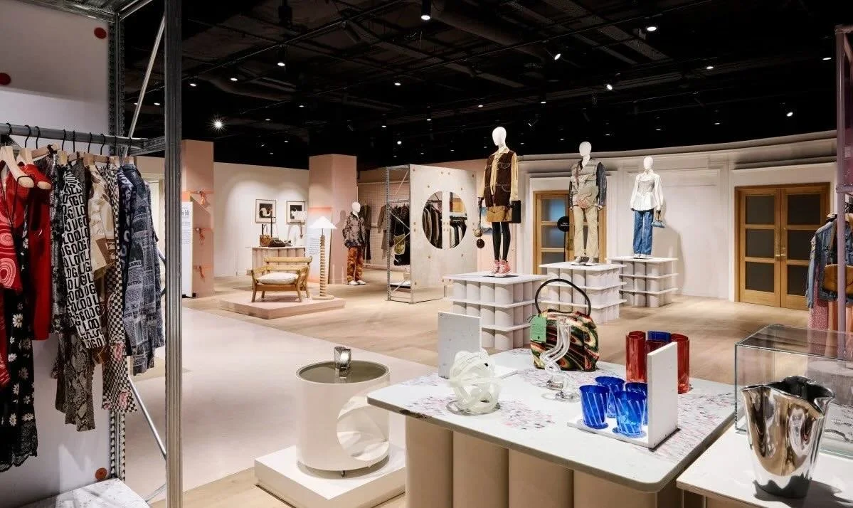 Retail clothing store with mannequins displaying outfits, racks of clothes, decorative items, and glassware on display tables.