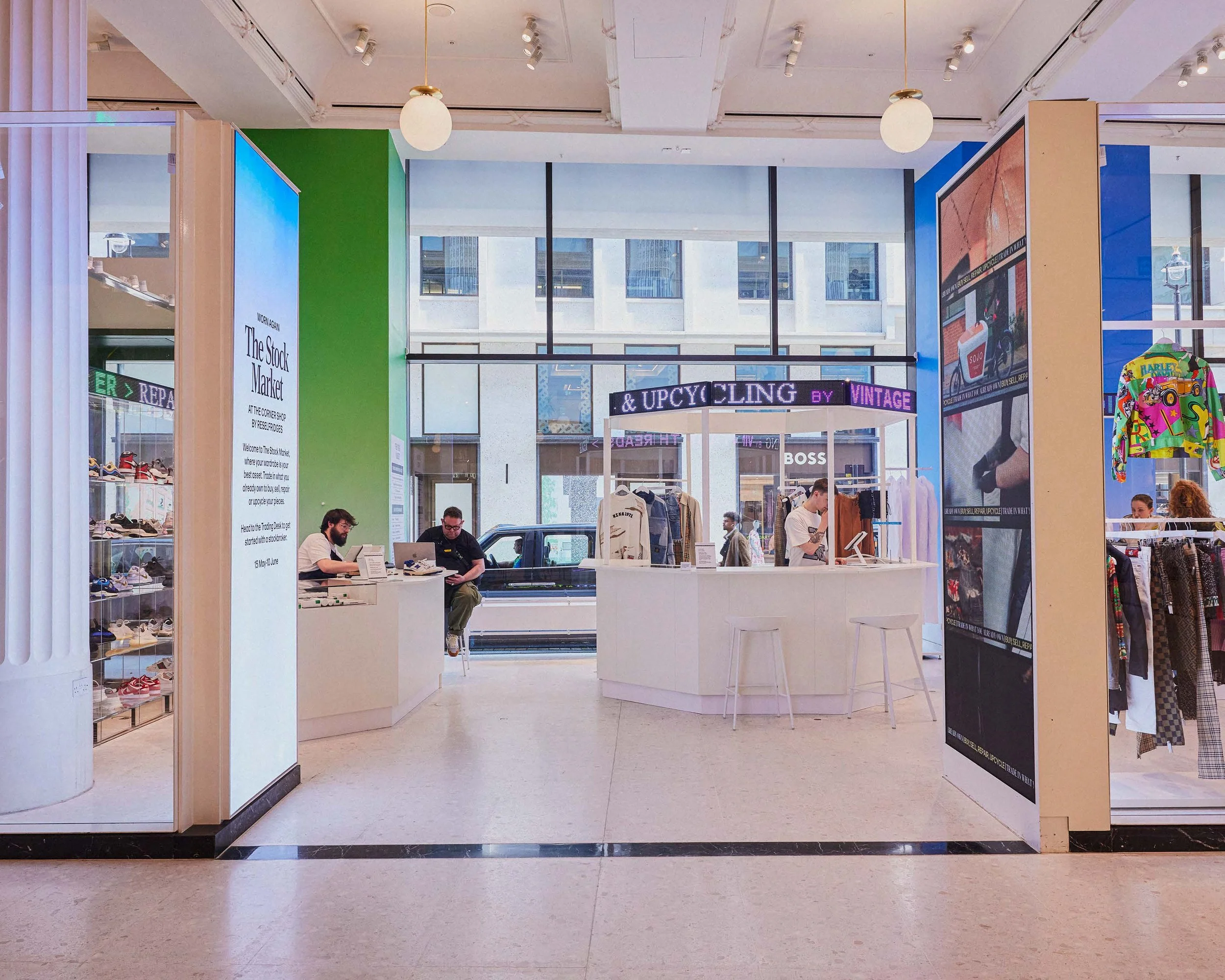 Interior view of a retail store with clothing racks, a customer service counter, colorful walls, large windows showing city buildings outside, and people shopping.