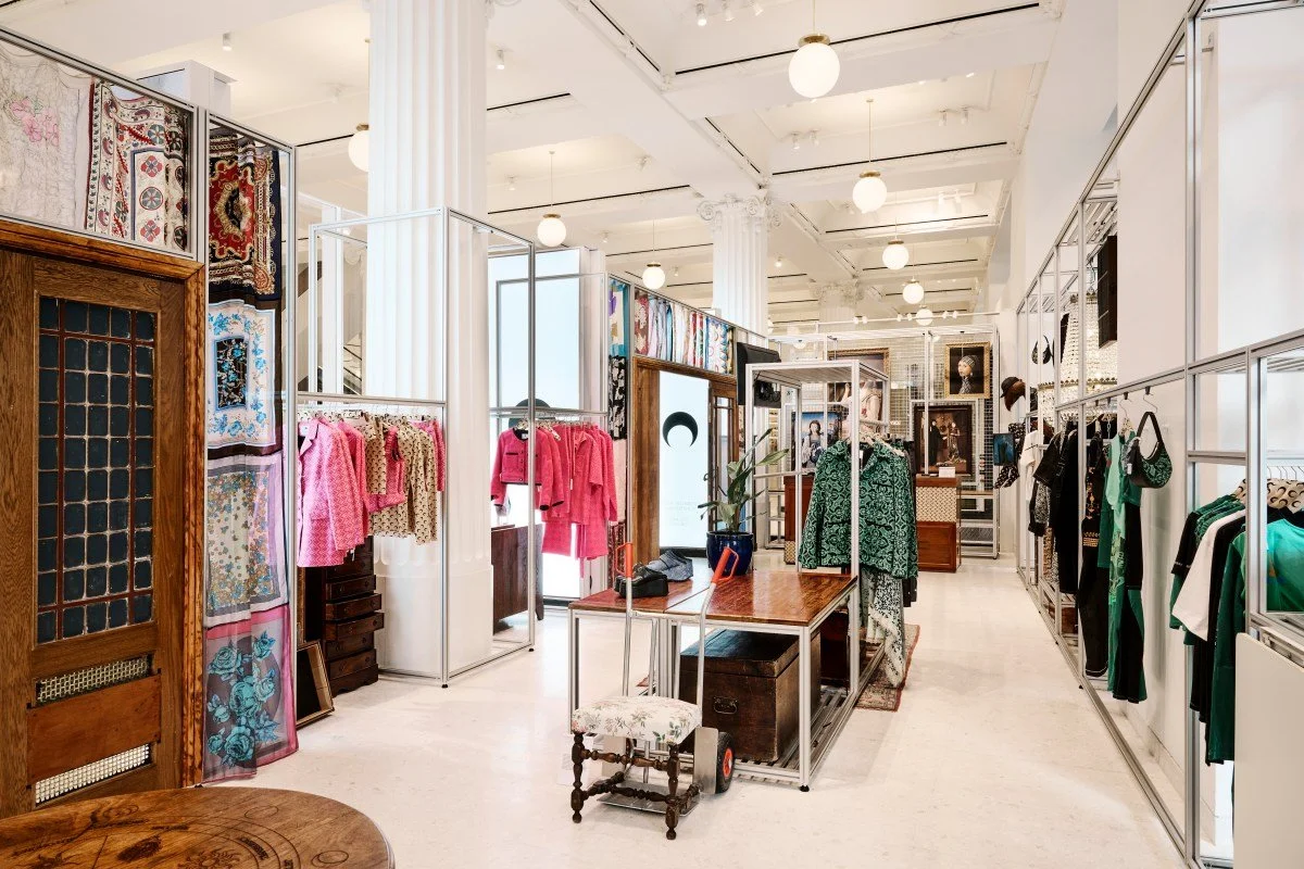 Interior of a boutique clothing store displaying various garments including pink jackets and green patterned coats on racks, with decorative wall hangings and framed artwork in the background, and a central wooden table with accessories and a small upholstered stool.