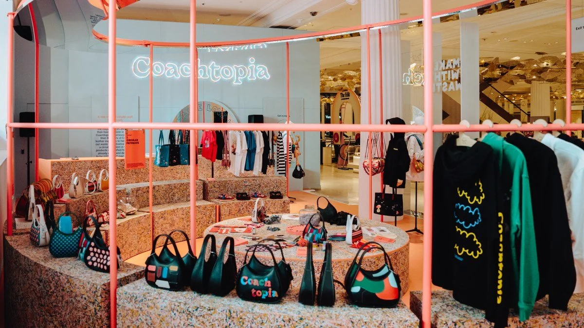 A retail store display of handbags, clothing, and accessories inside a shopping mall, with a sign saying "Coachtopia" in the background.