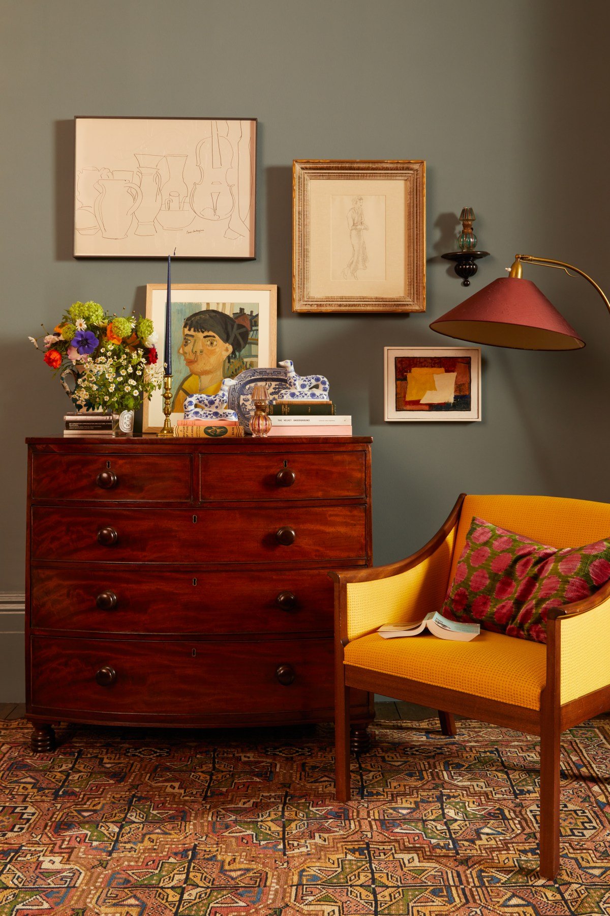 A decorated interior with a wooden dresser topped with flowers, artwork, a ceramic decorative object, and books. Above the dresser are framed artworks on a grey wall. To the right, a yellow armchair with a decorative pillow and a book, and a red/shaded floor lamp beside it.