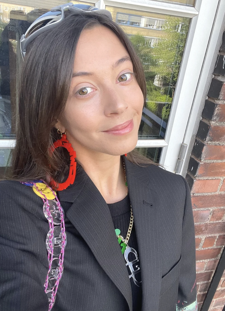 A woman with long brown hair wearing a black blazer and red earrings, standing outside near a glass window and brick wall, smiling at the camera during daytime.