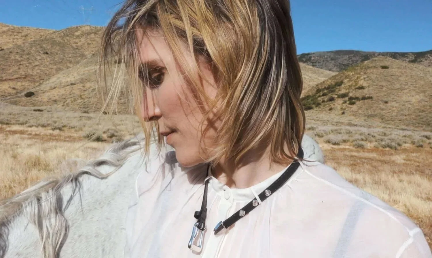 A woman with shoulder-length blonde hair looking down and to the side, standing outdoors in a dry, hilly landscape with a clear blue sky.