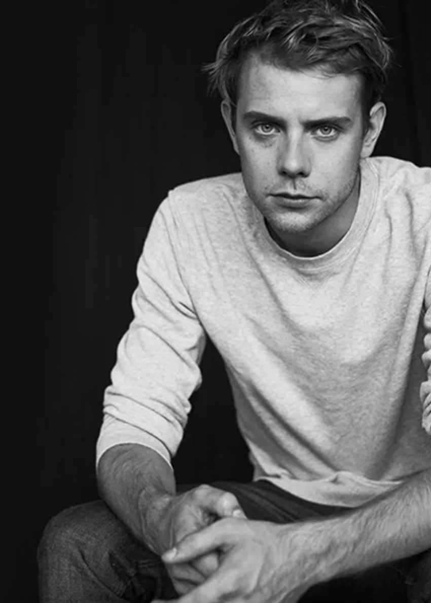 A black and white portrait of a young man with short, styled hair, wearing a light-colored long-sleeve shirt, sitting with hands clasped, and looking directly at the camera with a serious expression.