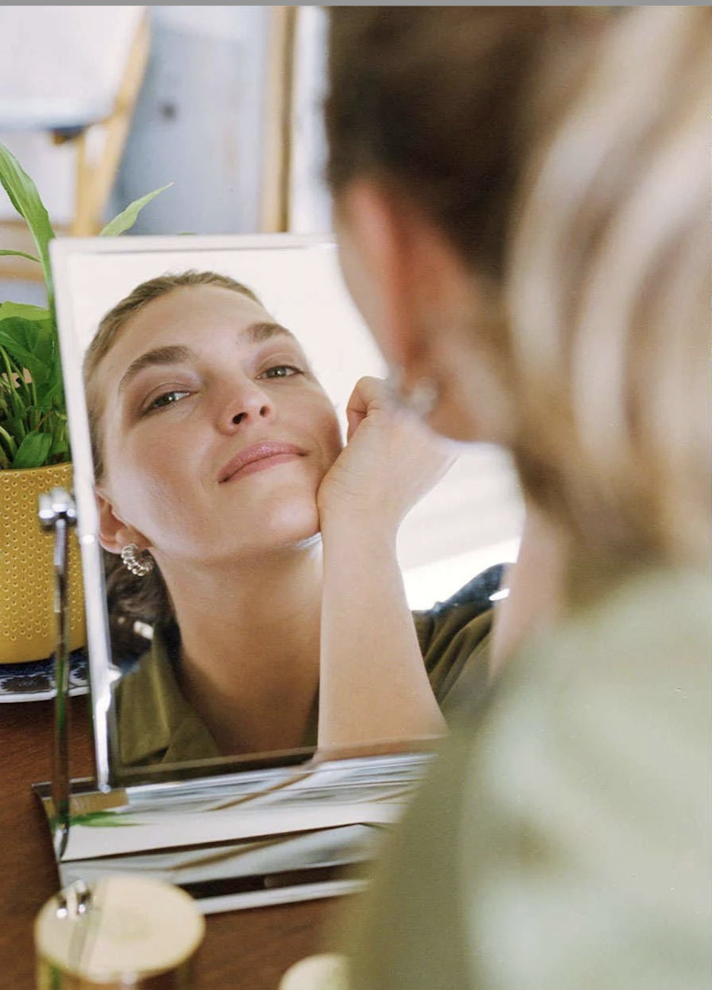 Woman looking at herself in a mirror, smiling gently, with her hand resting on her face.