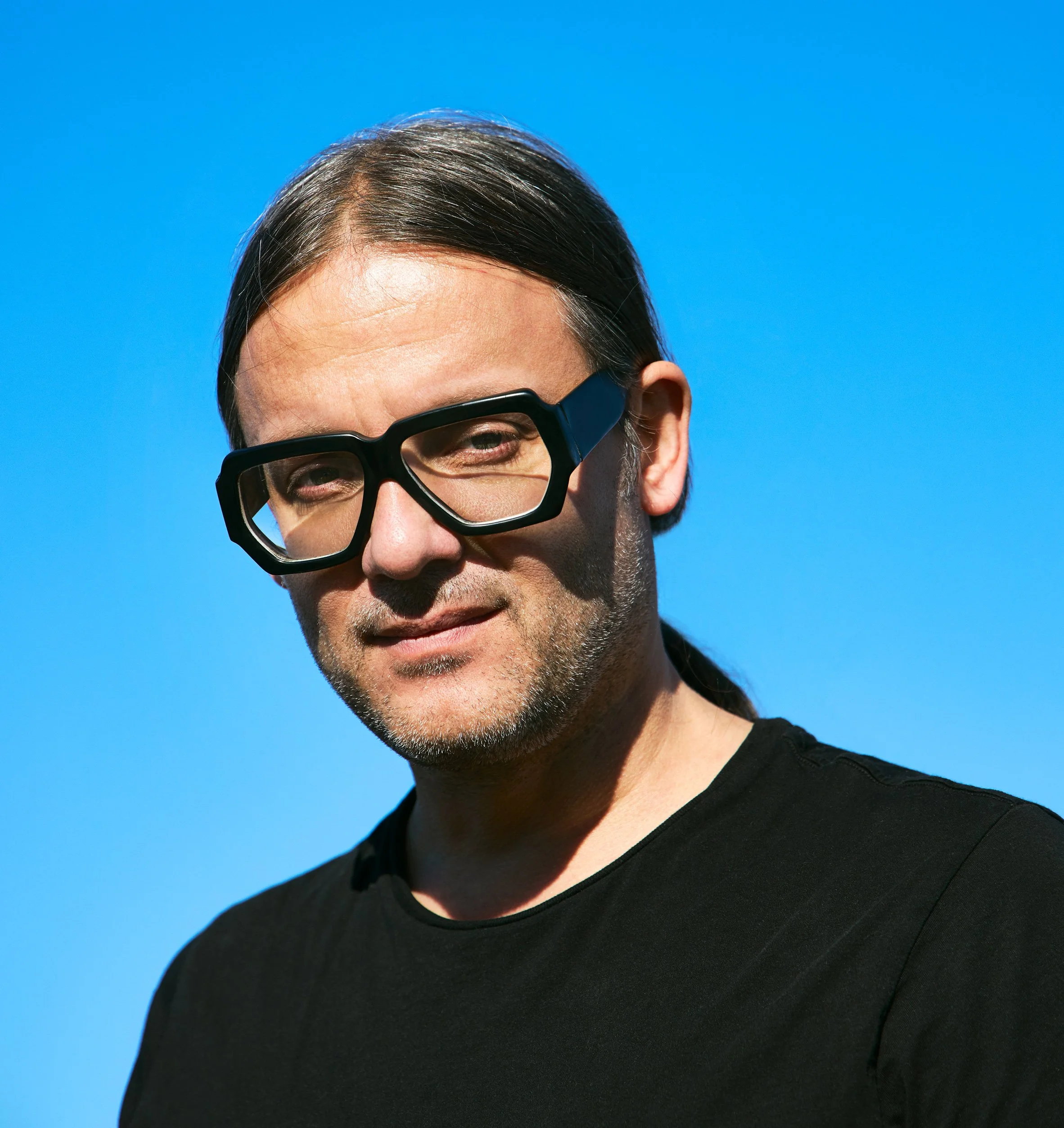 Close-up of a person wearing large black glasses, dark hair, and a black shirt, with a bright blue background.