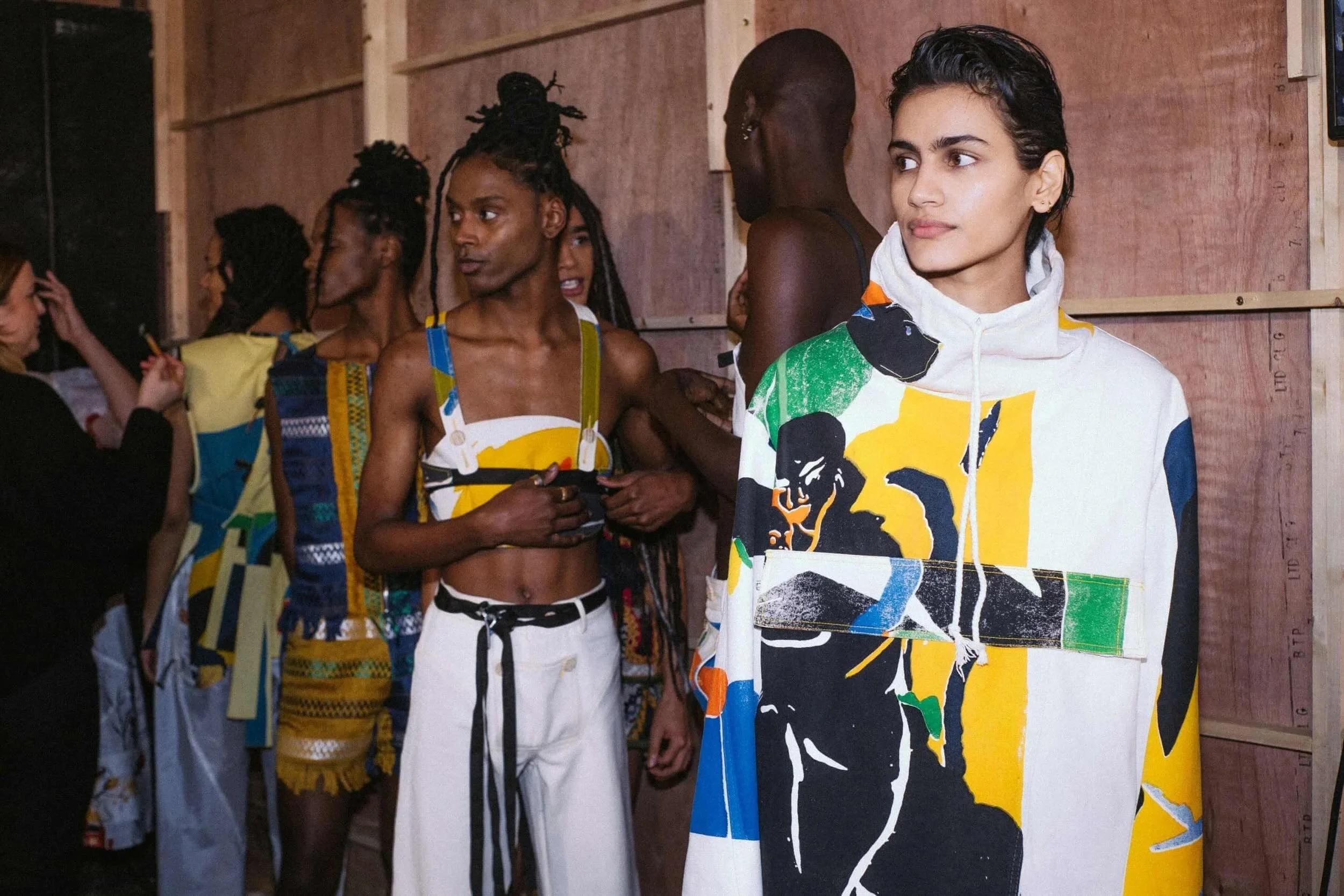 Group of diverse people, including a woman in a colorful patterned hoodie and others in athletic wear, standing against a wooden background. One person is holding a phone, and they appear to be backstage or preparing for an event.