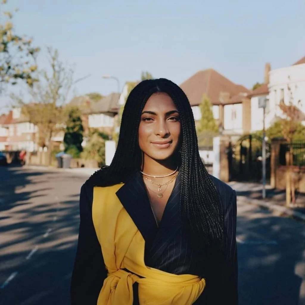 A woman with long braided black hair standing outdoors in front of residential houses during daytime.