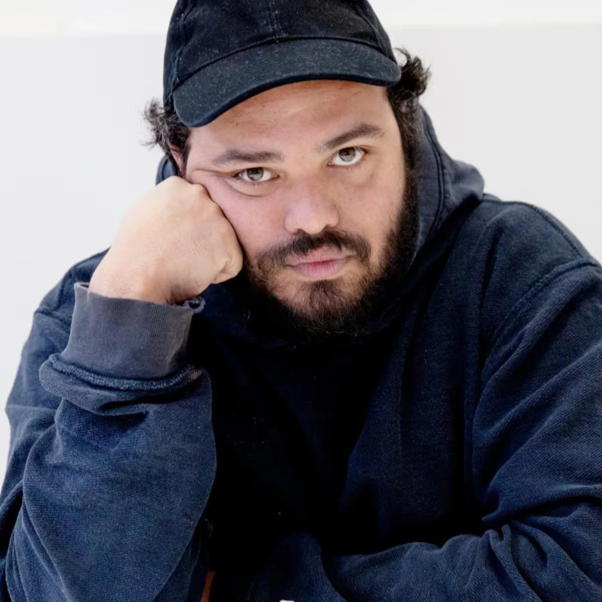 A man with a beard and mustache wearing a black baseball cap and a dark hoodie, resting his head on his hand, looking at the camera with a neutral expression.