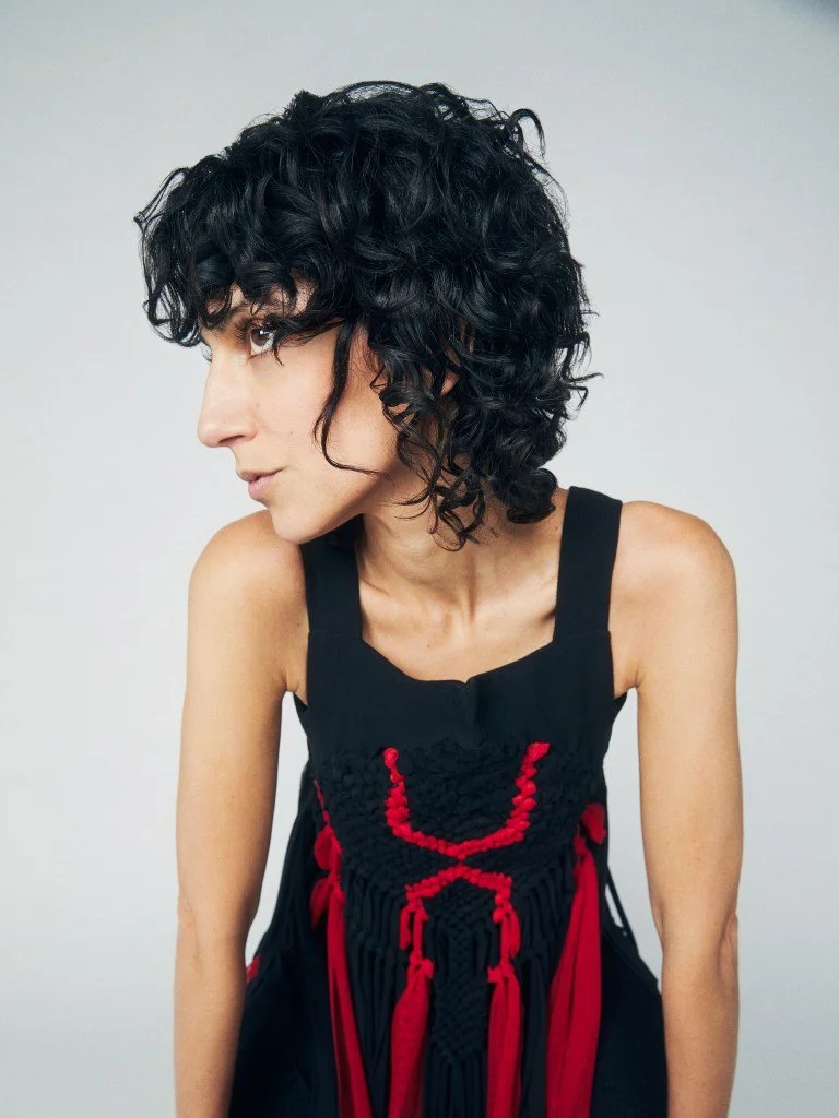 A woman with curly black hair wearing a black dress with red and black macrame details.
