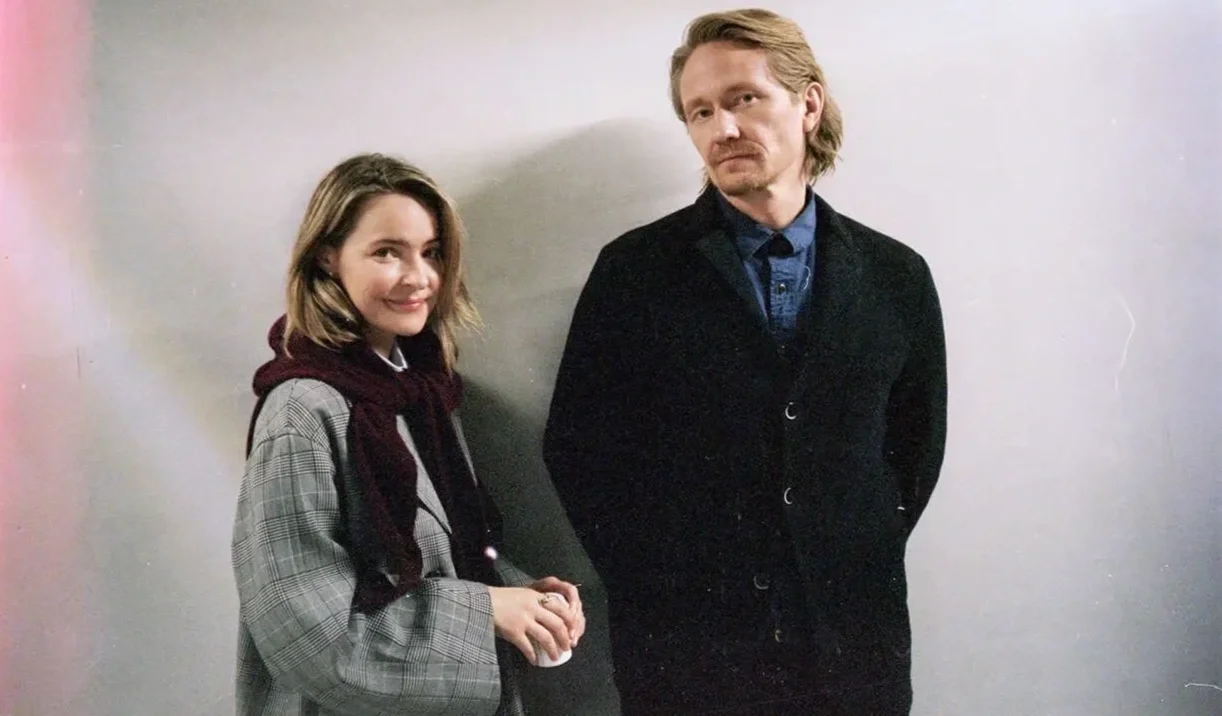 A young woman smiling and a man with a serious expression standing against a light-colored wall.