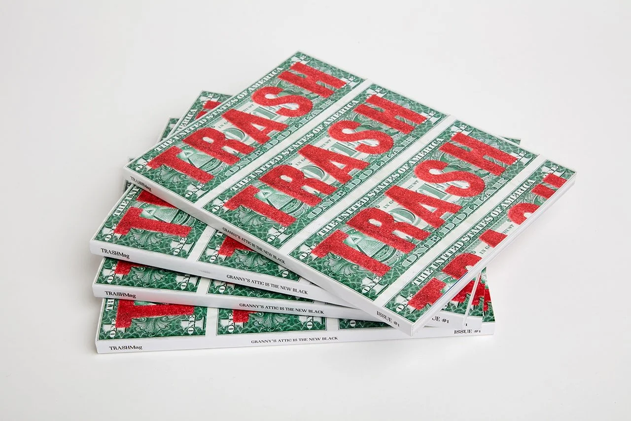 Stack of magazines with a dollar bill design cover, the word 'TRASH' printed in large red letters.