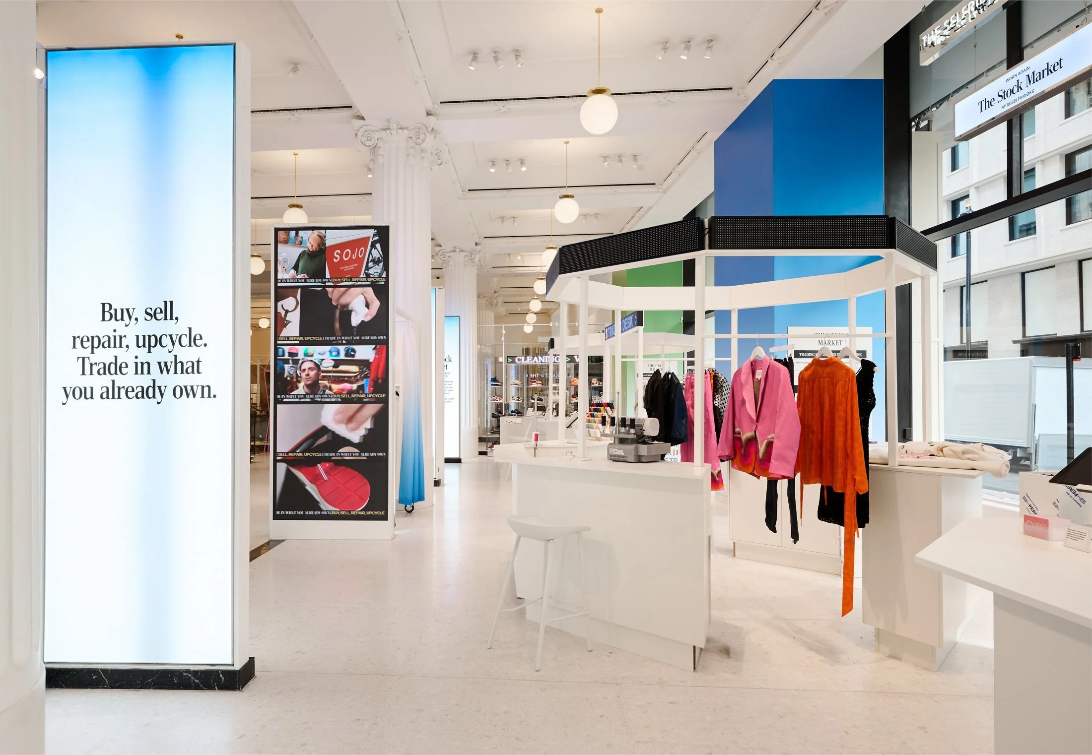 Interior of a modern clothing store with display tables and racks of clothing, large digital screens with advertisements, and informational signs, bright lighting, tall windows, and architectural columns.