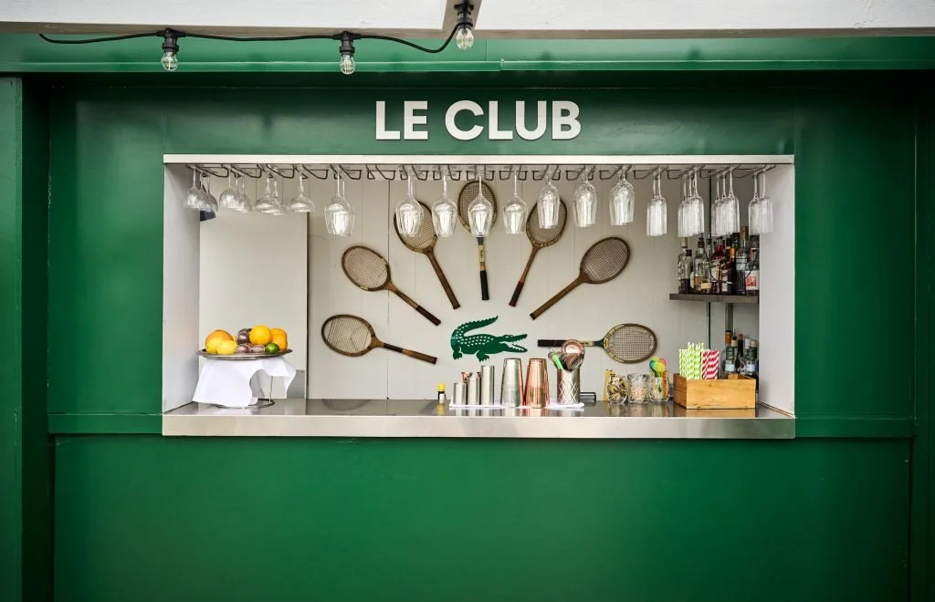 Bar with green front and white back wall, featuring hanging wine glasses, tennis racquets on the wall, a crocodile logo, bottles on a shelf, and a fruit bowl.