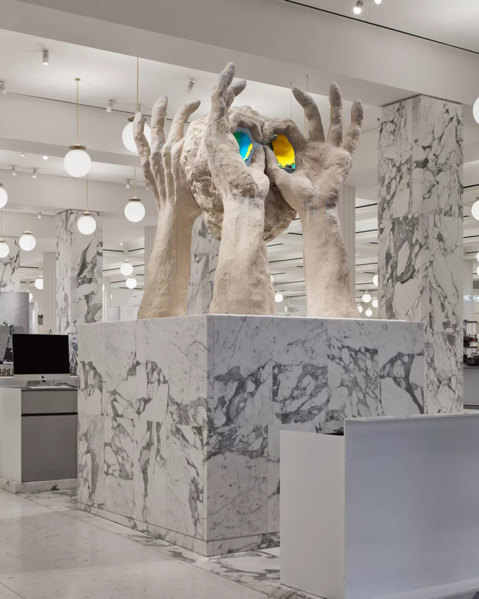A marble sculpture of multiple hands stacked and holding a colorful, abstract object in a modern, well-lit interior space.