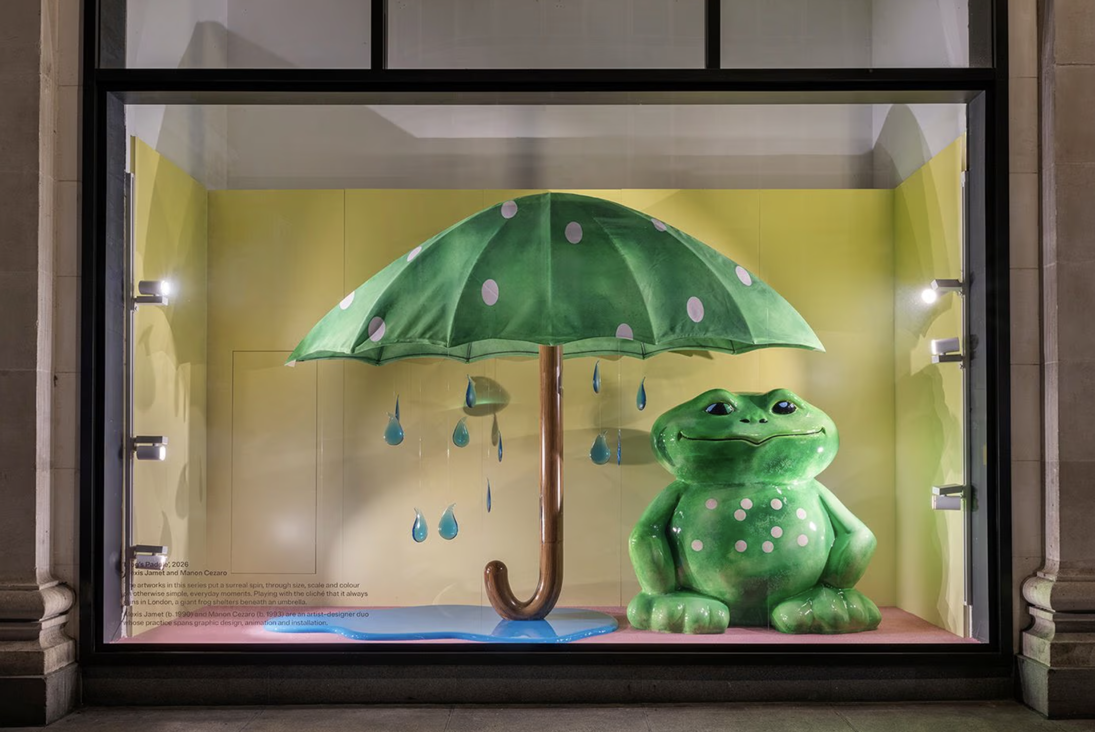 Display window showing a green frog-shaped sculpture holding a green umbrella with white polka dots, with raindrop decorations hanging from the umbrella.