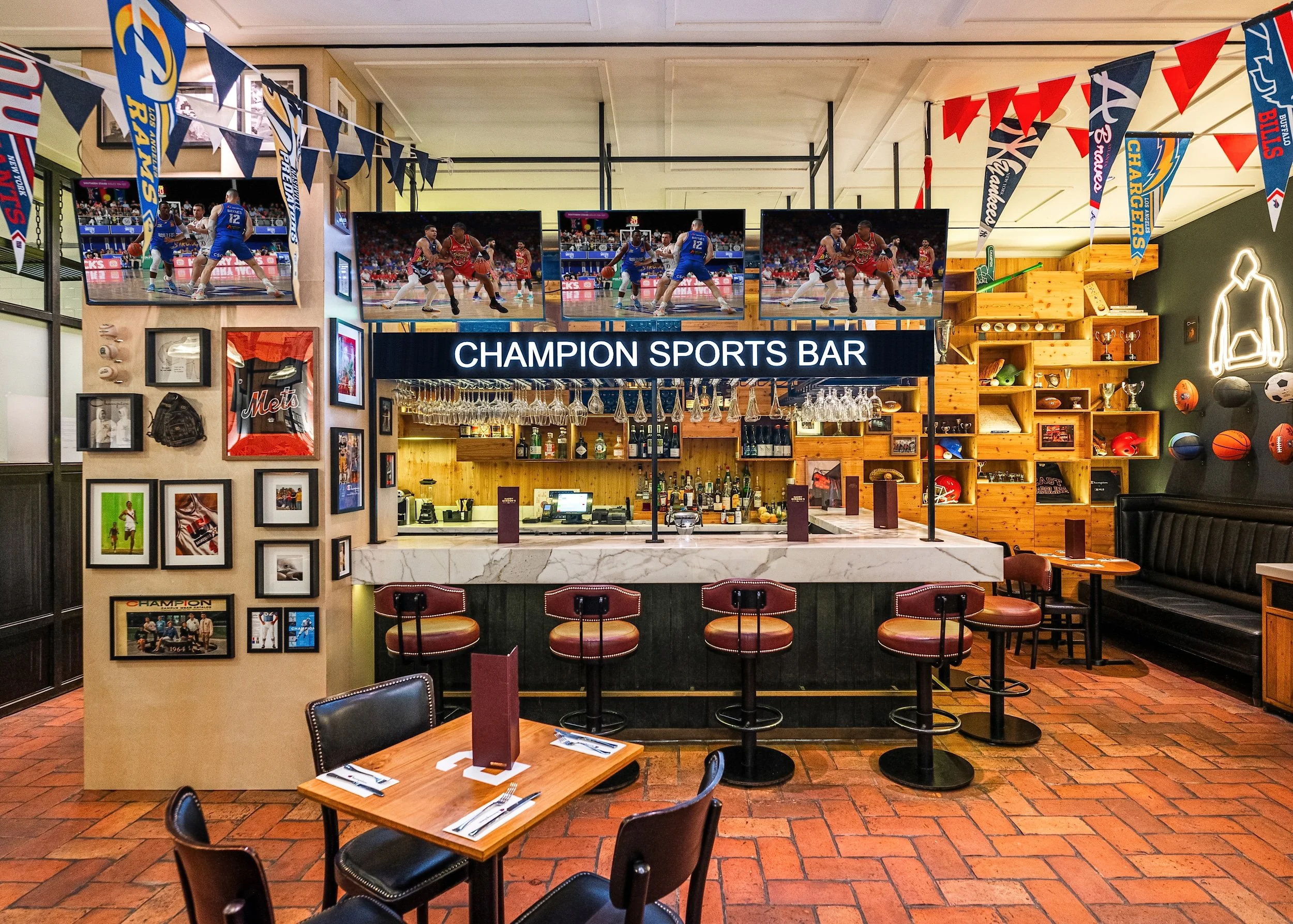 Interior of a sports bar decorated with sports memorabilia, including pennants and framed photos, with multiple flat-screen TVs displaying basketball games, a bar area with bottles and glasses, and tables and chairs for patrons.