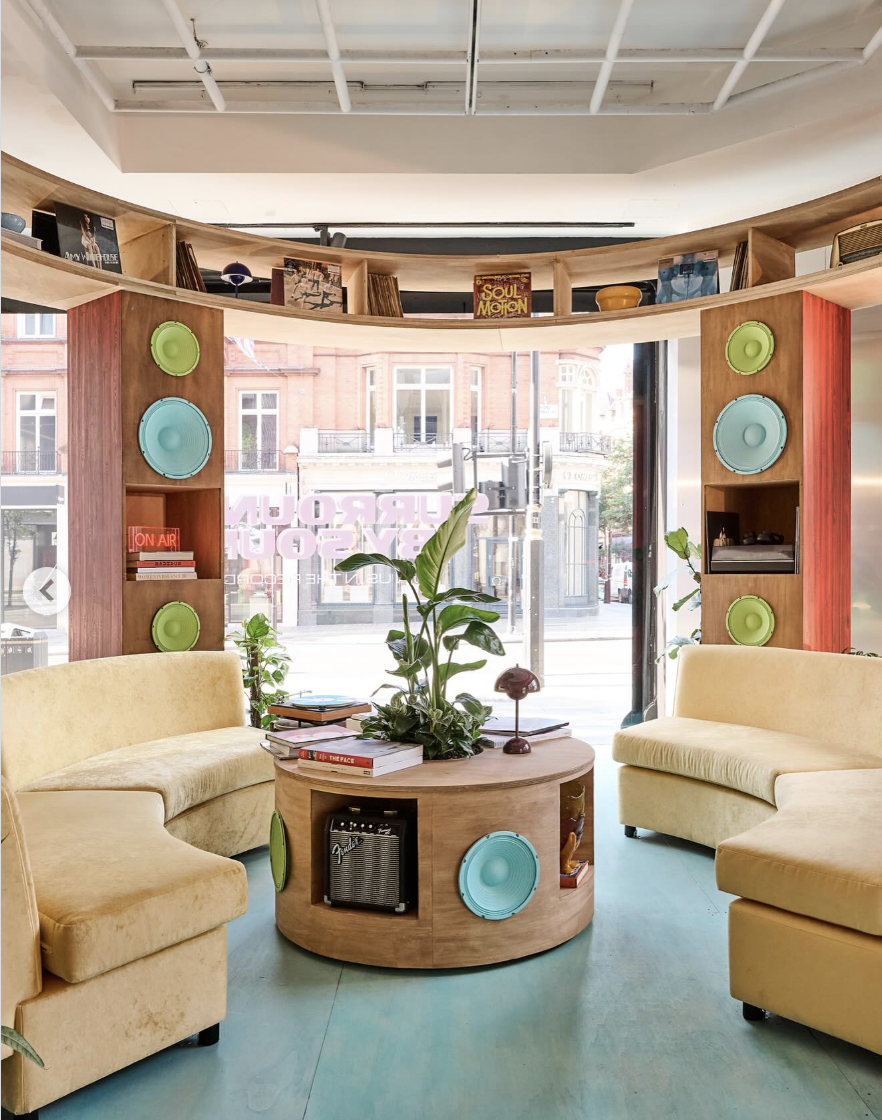 Interior of a cozy lounge with yellow sofas, a round wooden coffee table, and built-in shelves with colorful speakers and books, large windows with street view.
