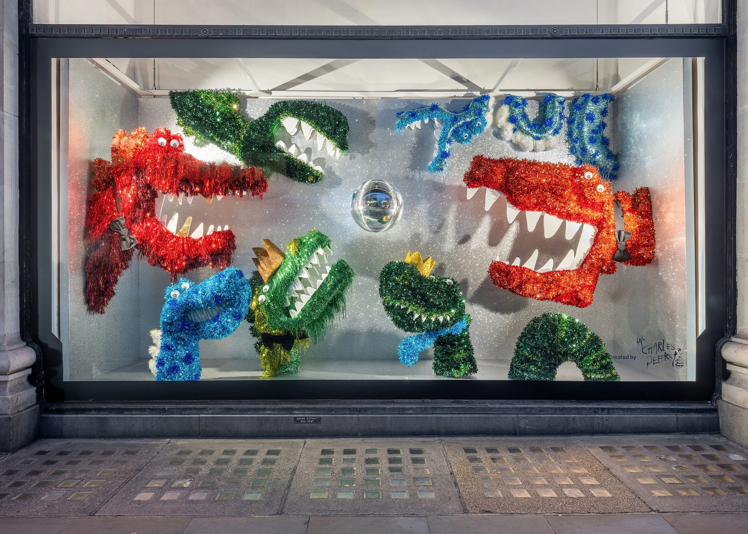 Holiday window display with colorful, glittery dinosaur decorations, including red and blue T-Rex and a blue pterosaur, with googly eyes and bow ties, set against a glittery silver background.