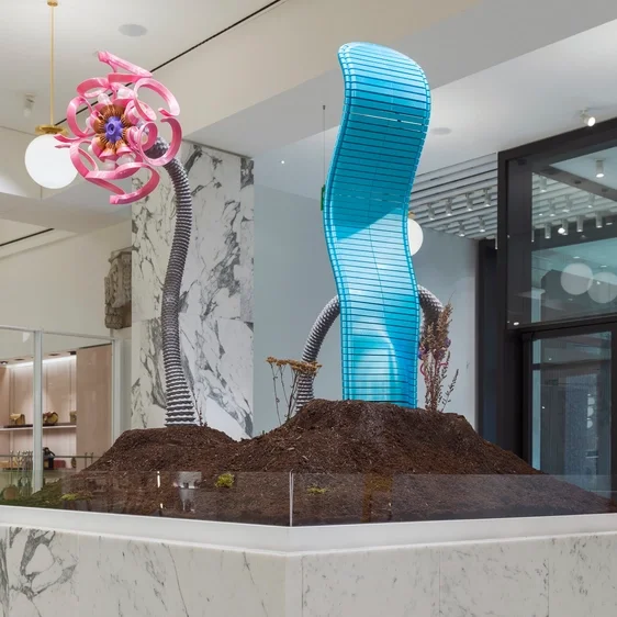 Indoor art installation with colorful sculptural elements resembling flowers and curved structures on a mound of soil inside a modern building.