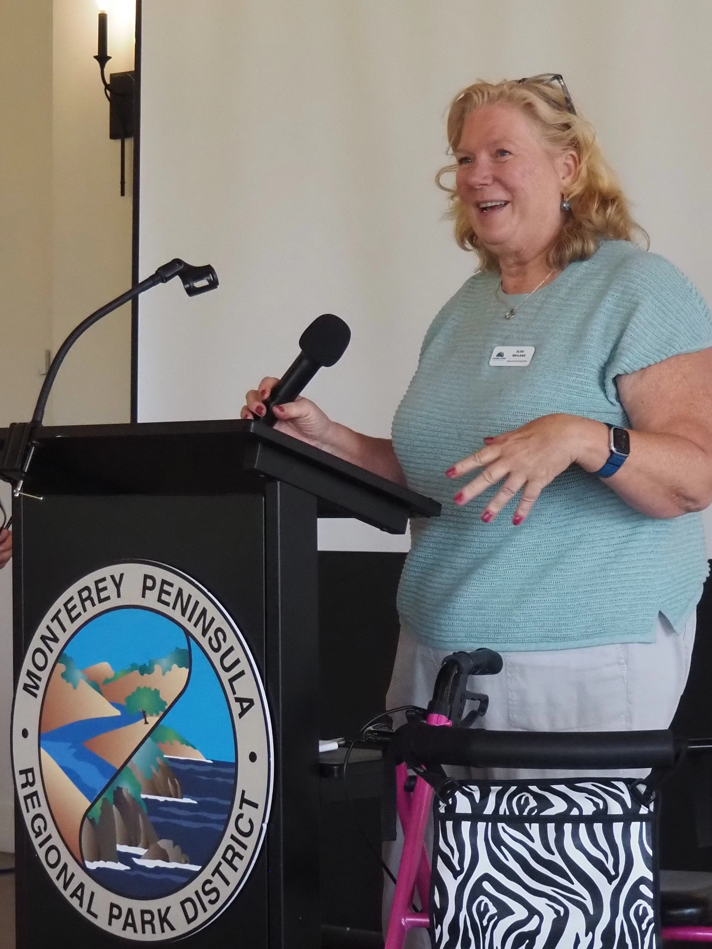 A woman speaking at a podium with a National Park District emblem, holding a microphone, standing in front of a beige wall, with a black wall sconce to her left, wearing a light green short-sleeved shirt, glasses on her head, earrings, and a name tag.