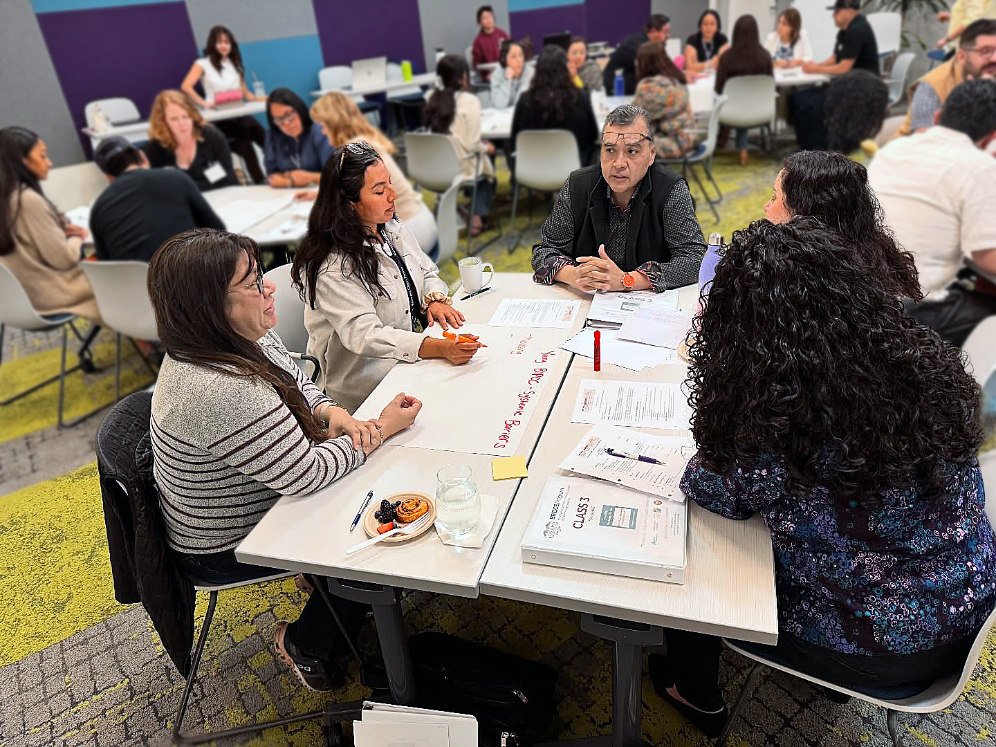Group of diverse adults participating in a workshop , sitting around tables with papers, notebooks, and coffee mugs.