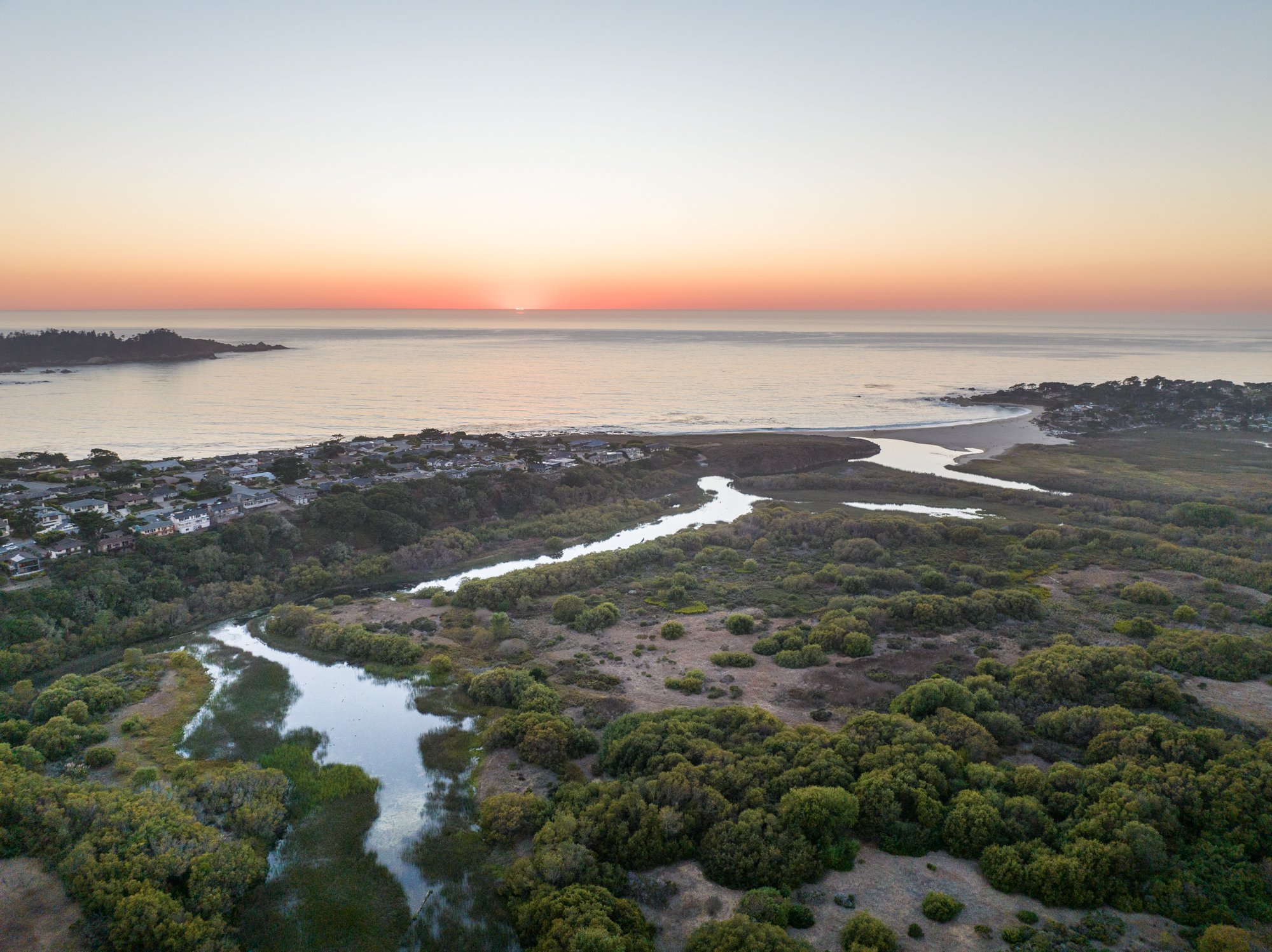 An aerial view of a coastal landscape at sunset, showing a beach, a small town with houses, winding river, and lush green vegetation.