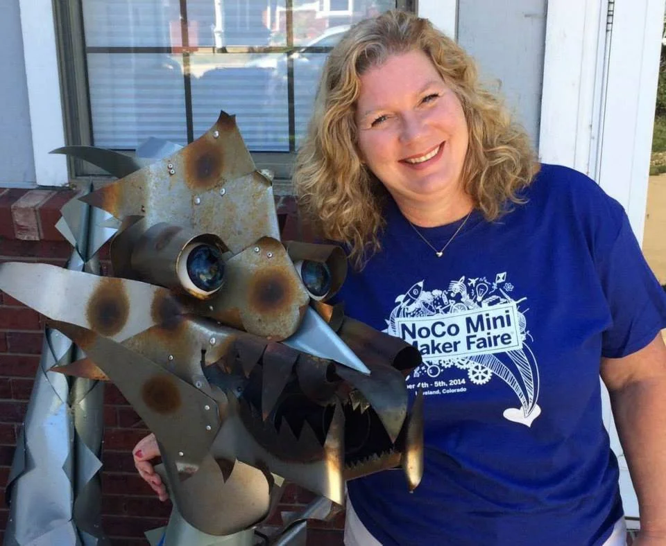 Elise with curly blonde hair, smiling and wearing a blue t-shirt that reads 'NoCo Mini Maker Faire,' stands next to a metal sculpture of a mythical creature outside a building with a brick wall and window.