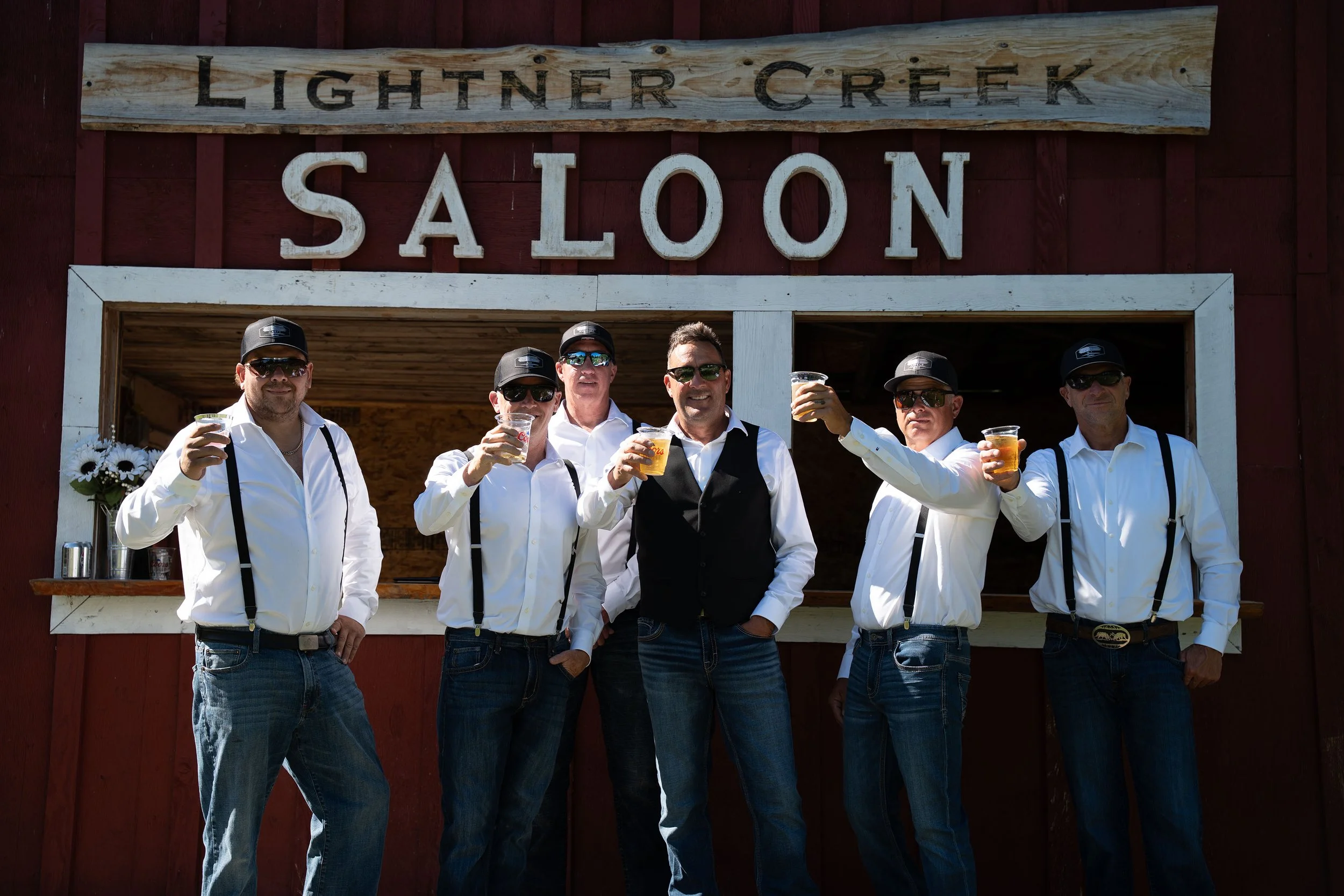 Group of six men dressed in white shirts, black suspenders, and sunglasses, holding drinks and standing in front of a rustic red building with a sign that reads 'Lightner Creek Saloon.'