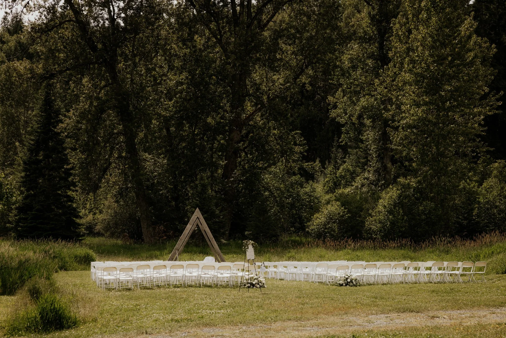 An outdoor wedding setup with rows of white chairs, a floral arrangement, and a triangle-shaped wooden arch in a grassy area surrounded by tall trees under a partly cloudy sky.