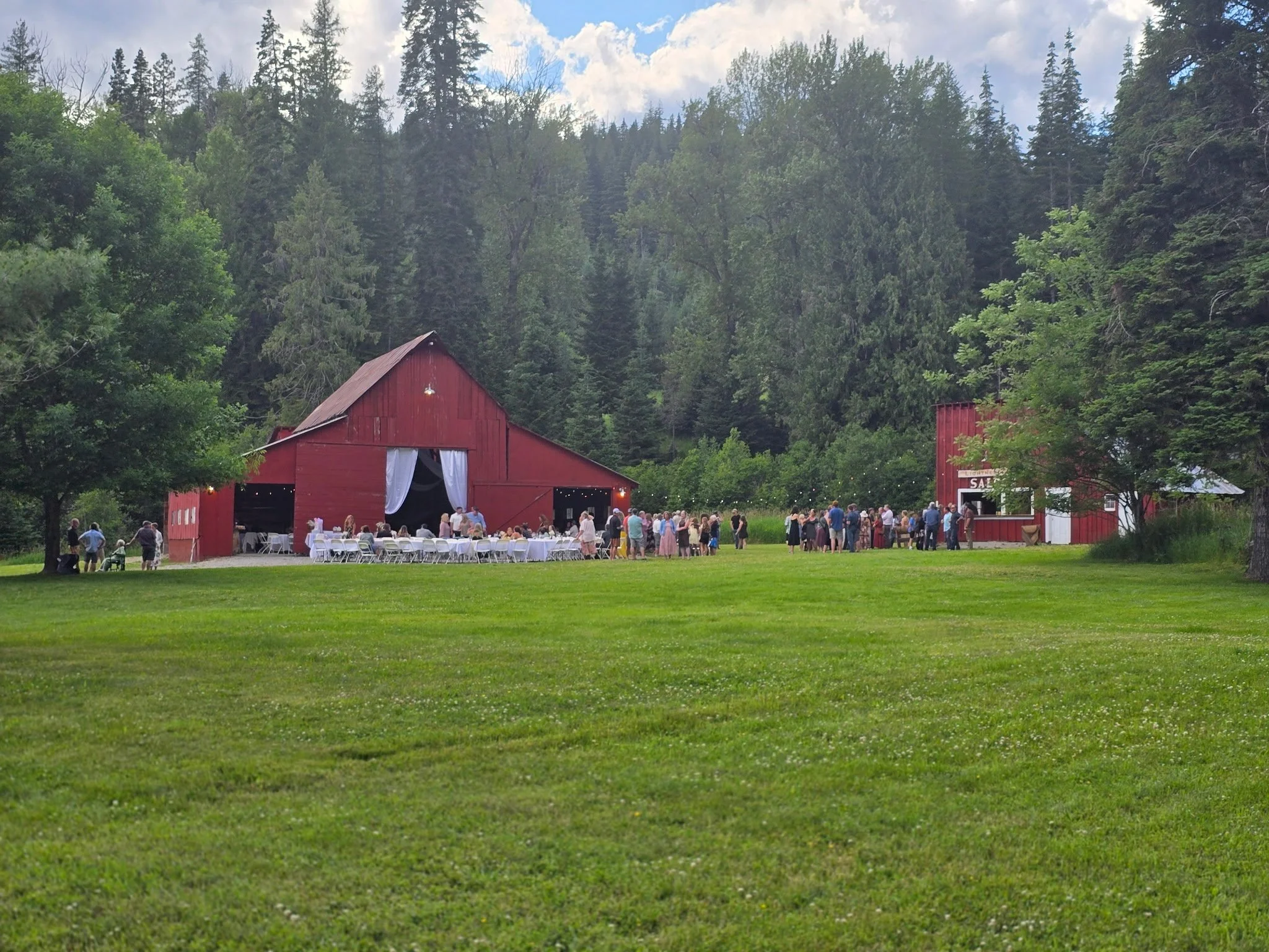 A large red barn-style building with white curtains on the open door, set in a lush green field. There are many people gathered outside, some seated at tables, others standing or walking, with a wooded forest and partly cloudy sky in the background.
