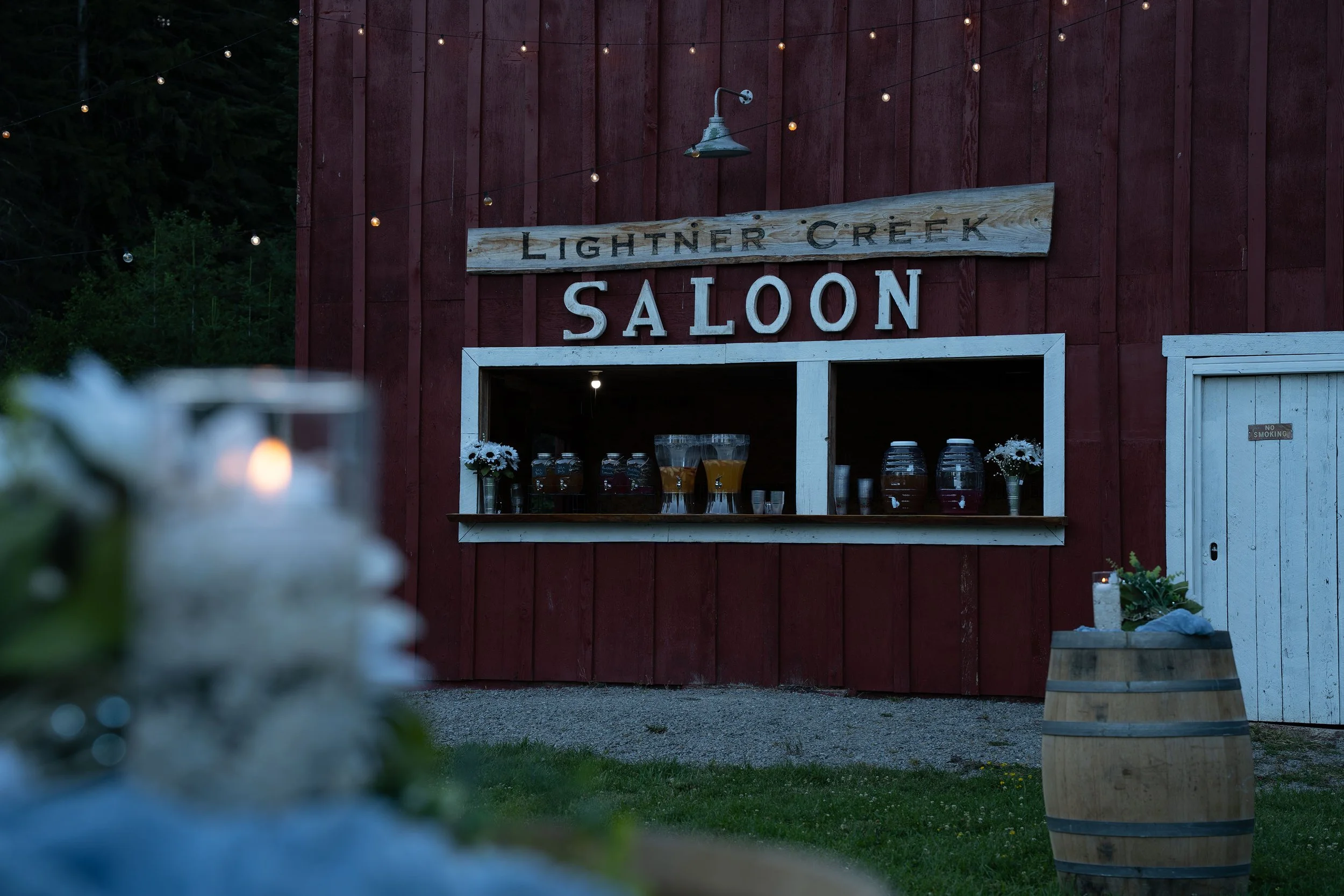 Wooden sign reading 'Lighter Creek Saloon' hangs on a red wooden building with string lights. Outdoor beverage station with jars of drinks is visible, and a wine barrel is in the foreground.