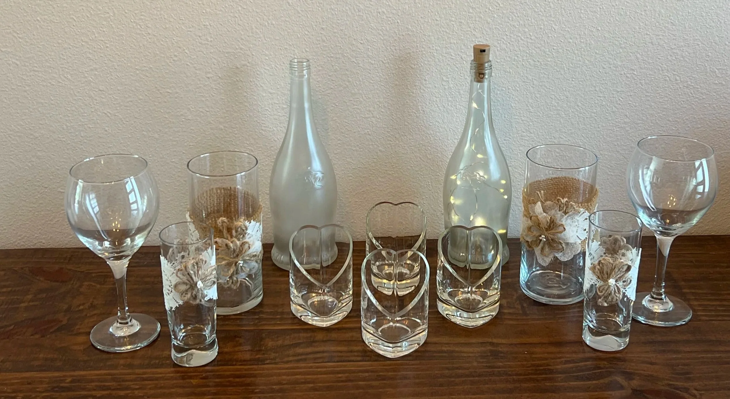 A display of various glassware including wine glasses, tumblers, and decorative bottles with fairy lights and ribbons, arranged on a wooden surface against a plain wall.