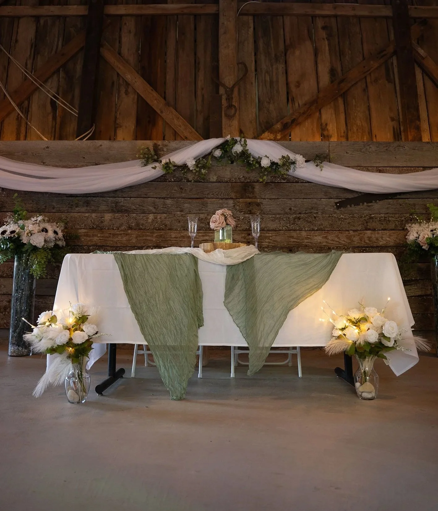 Wedding reception table setup with white tablecloth, green fabric drapes, floral arrangements, and candles, against a rustic wooden backdrop.