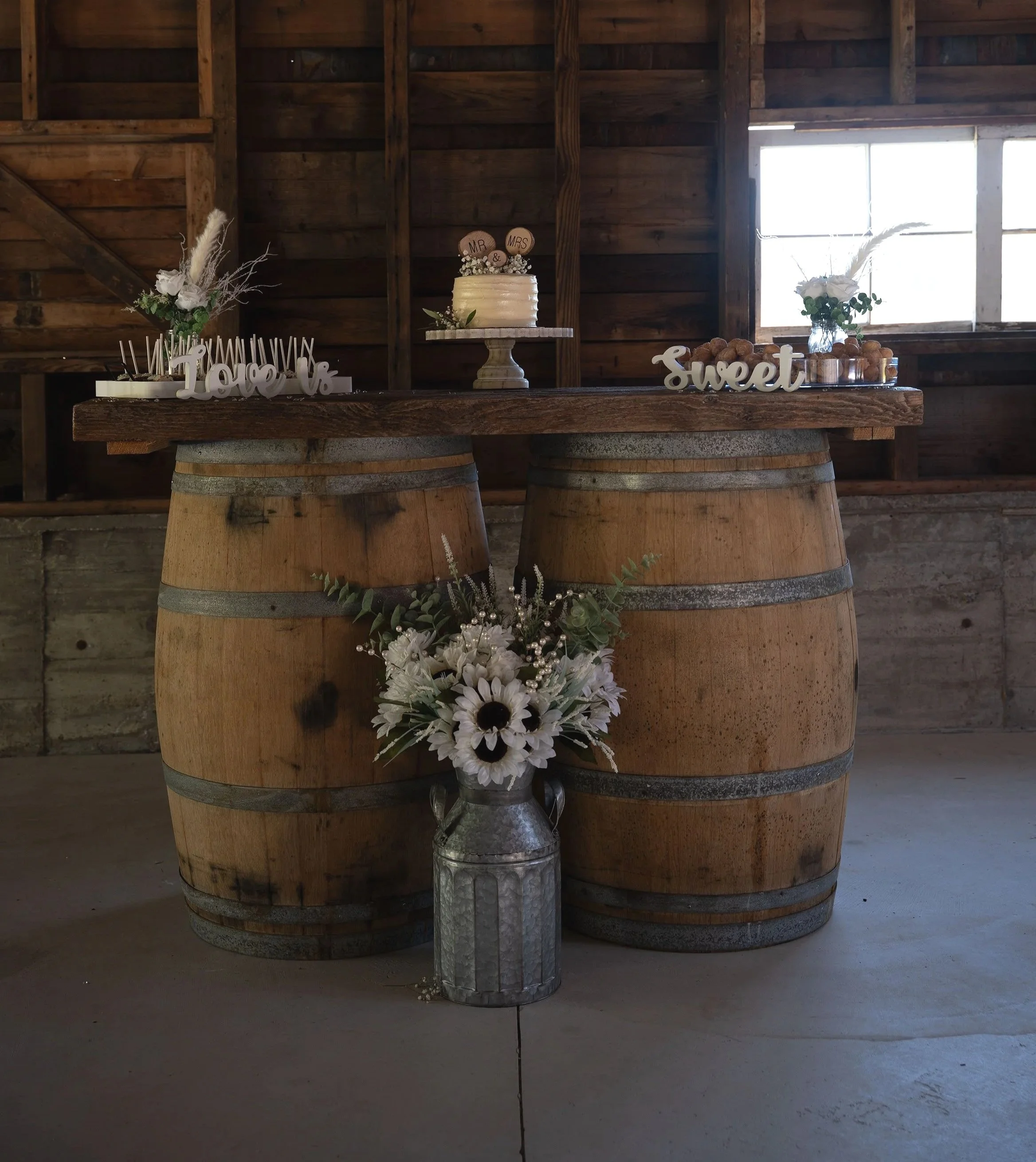 A rustic wedding display on a wooden table with a small white wedding cake topped with wooden wedding-themed decor, floral arrangements with white roses and greenery on either side, decorative words 'Love' and 'Sweet' on the table, and a metal wateri