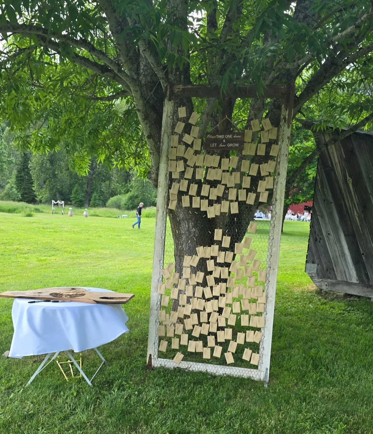 A tree with notes pinned to its trunk, surrounded by a rural landscape with green grass, a person walking, and a wooden barn.