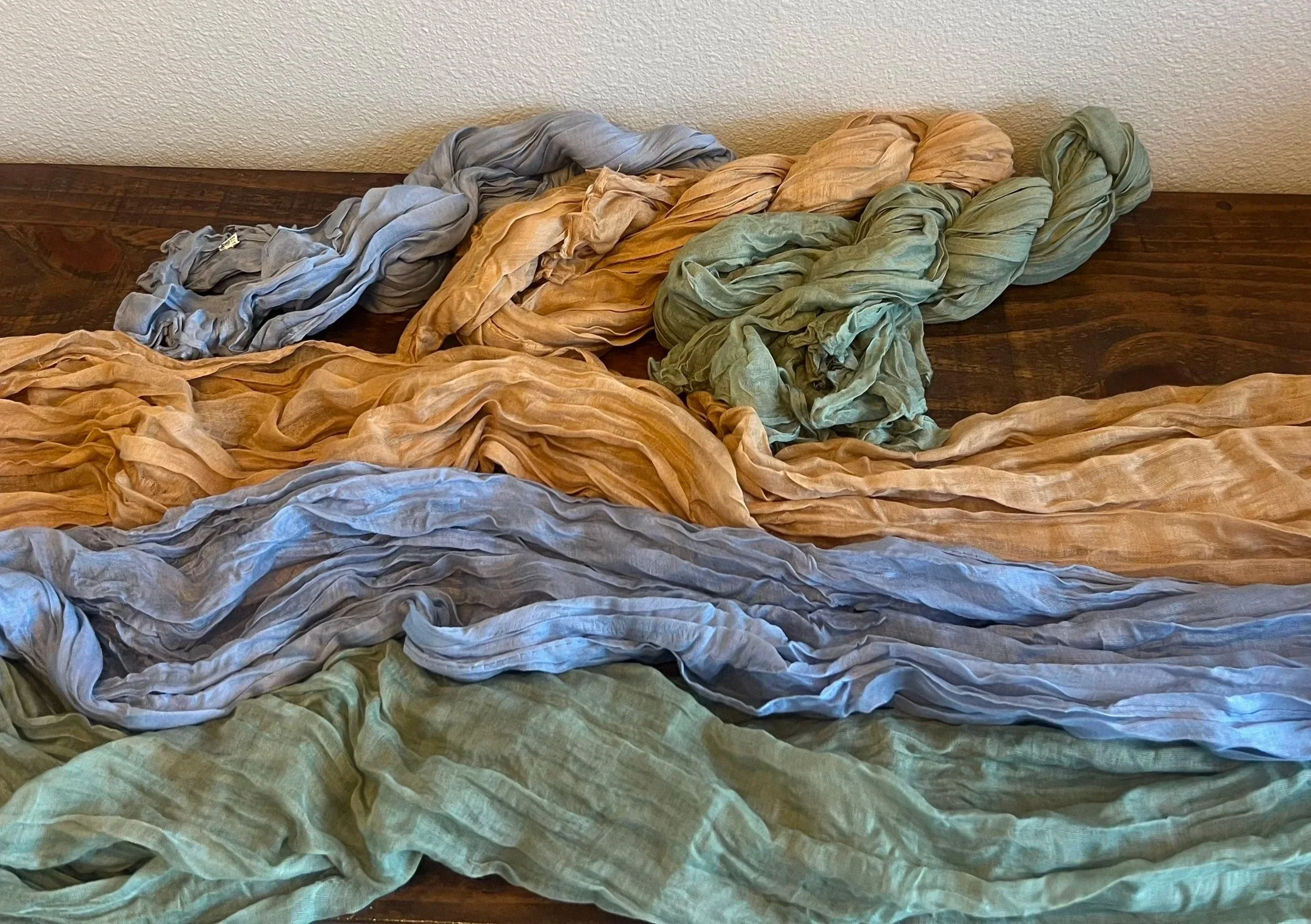Colorful crumpled fabric scarves in pastel shades of blue, yellow, green, and beige arranged on a dark wooden surface.