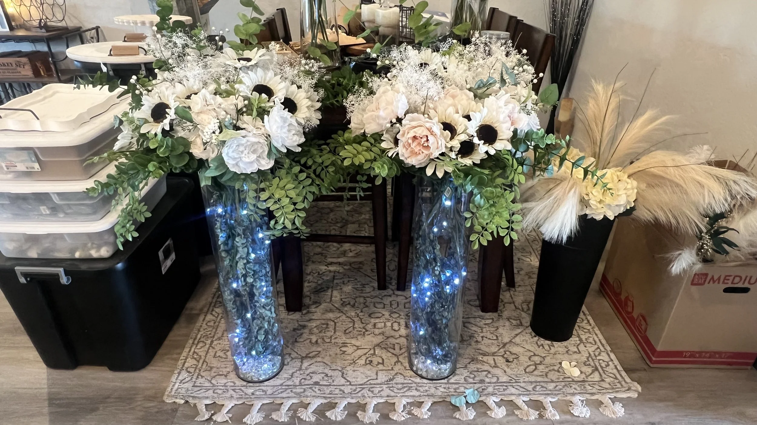 A floral arrangement on a dining table with white peonies, roses, anemones, greenery, and decorative pampas grass, flanked by tall glass vases with fairy lights inside.
