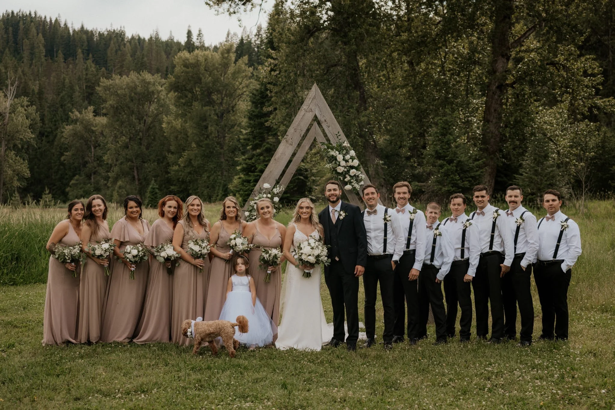 Group of people at a wedding outdoors, standing together on grass with trees in the background, a large wooden triangular arch decorated with flowers.