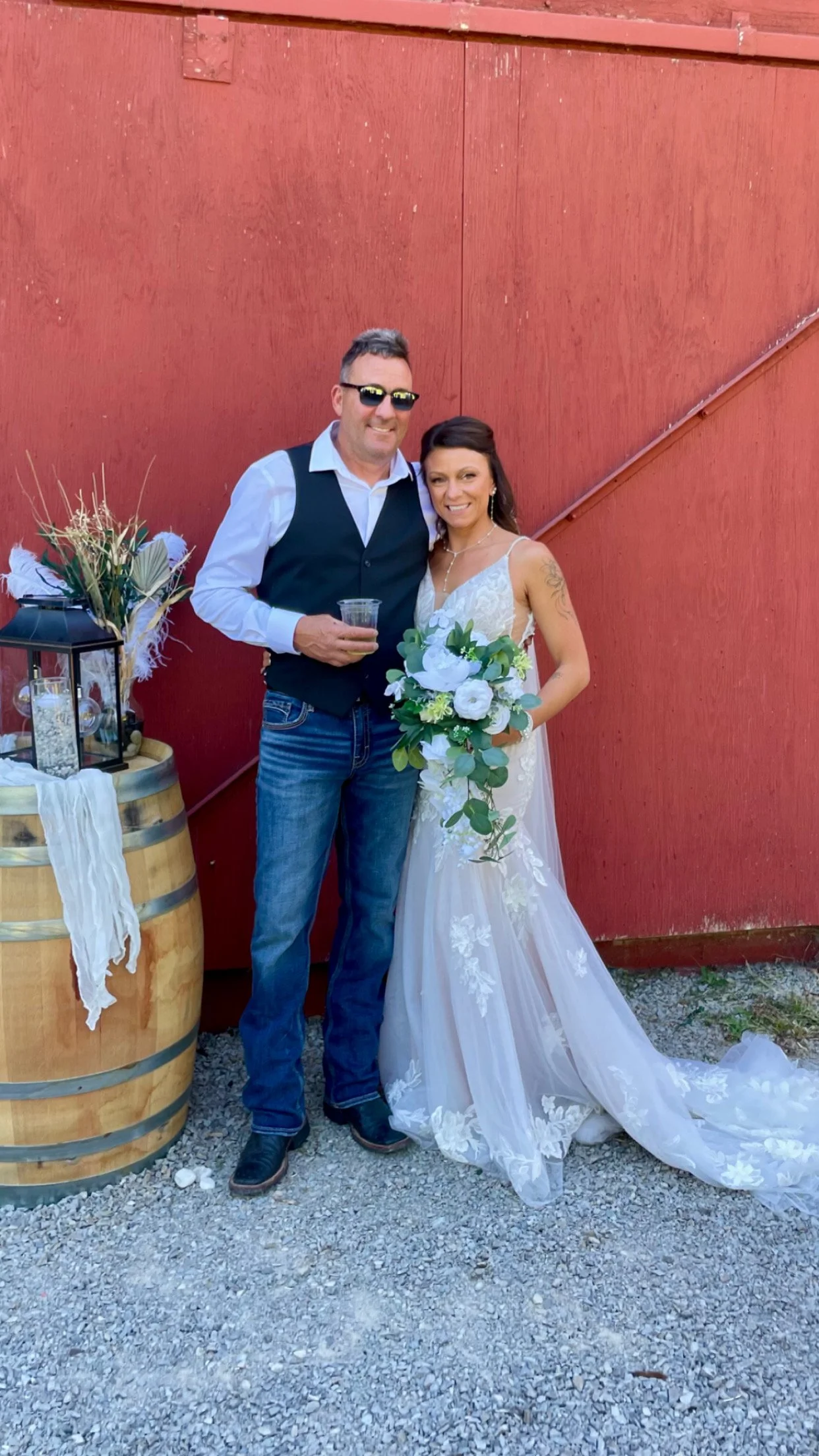 A man and woman in wedding attire standing in front of a red barn wall. The man is wearing sunglasses, a white shirt, a black vest, and jeans. The woman is holding a bouquet of white flowers and greenery, wearing a white lace wedding dress, with dark