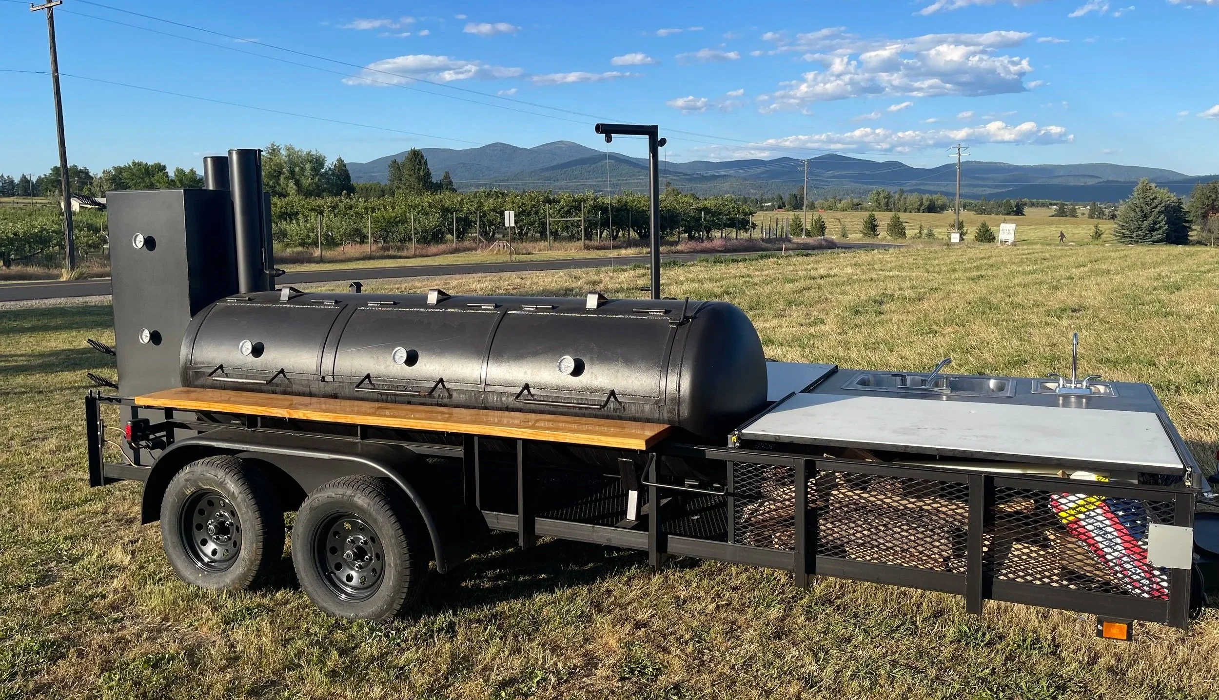 Black barbecue smoker on a trailer with a metal prep sink, set on a grassy field with vineyard and mountain landscape in the background.