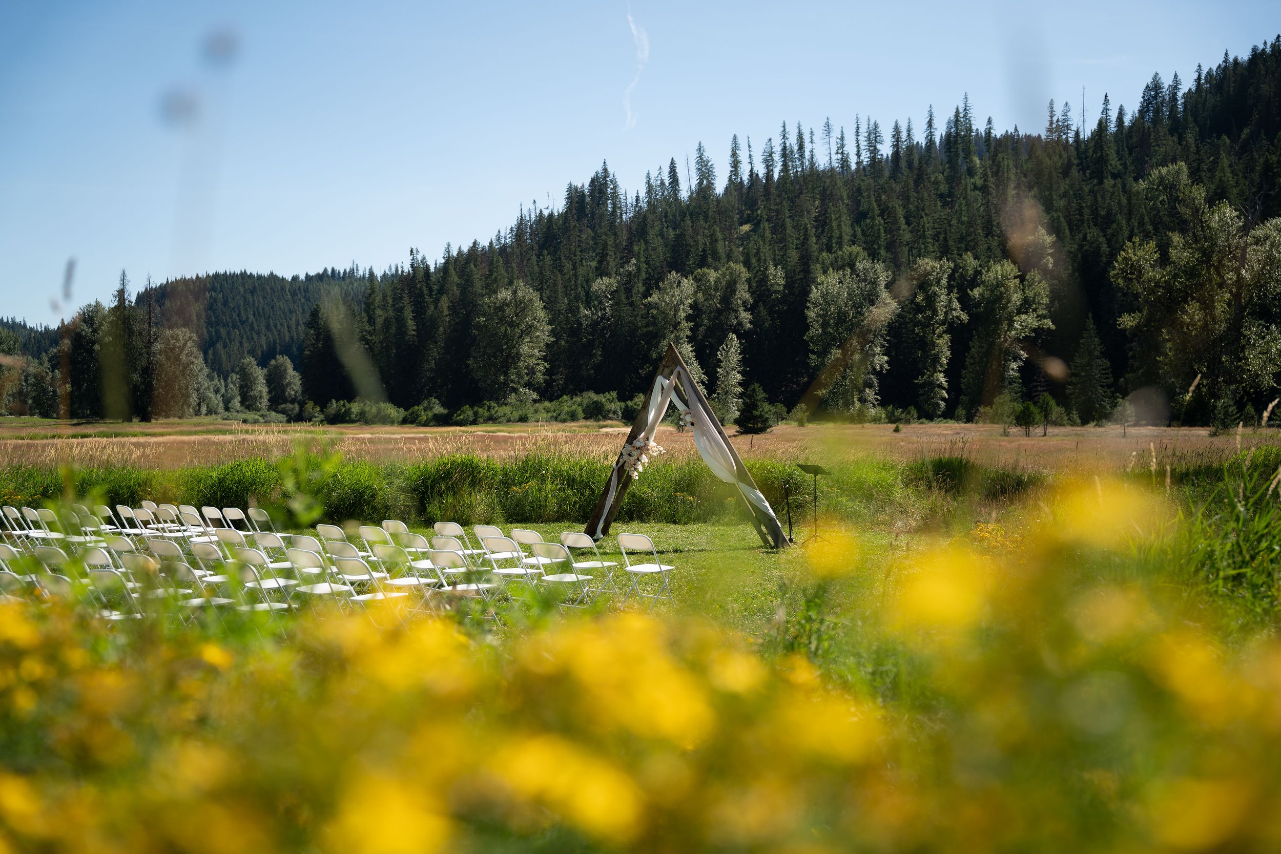 An outdoor wedding setup with white chairs arranged on a grassy field in front of a mountain and forest backdrop, with a decorative arch in the center.