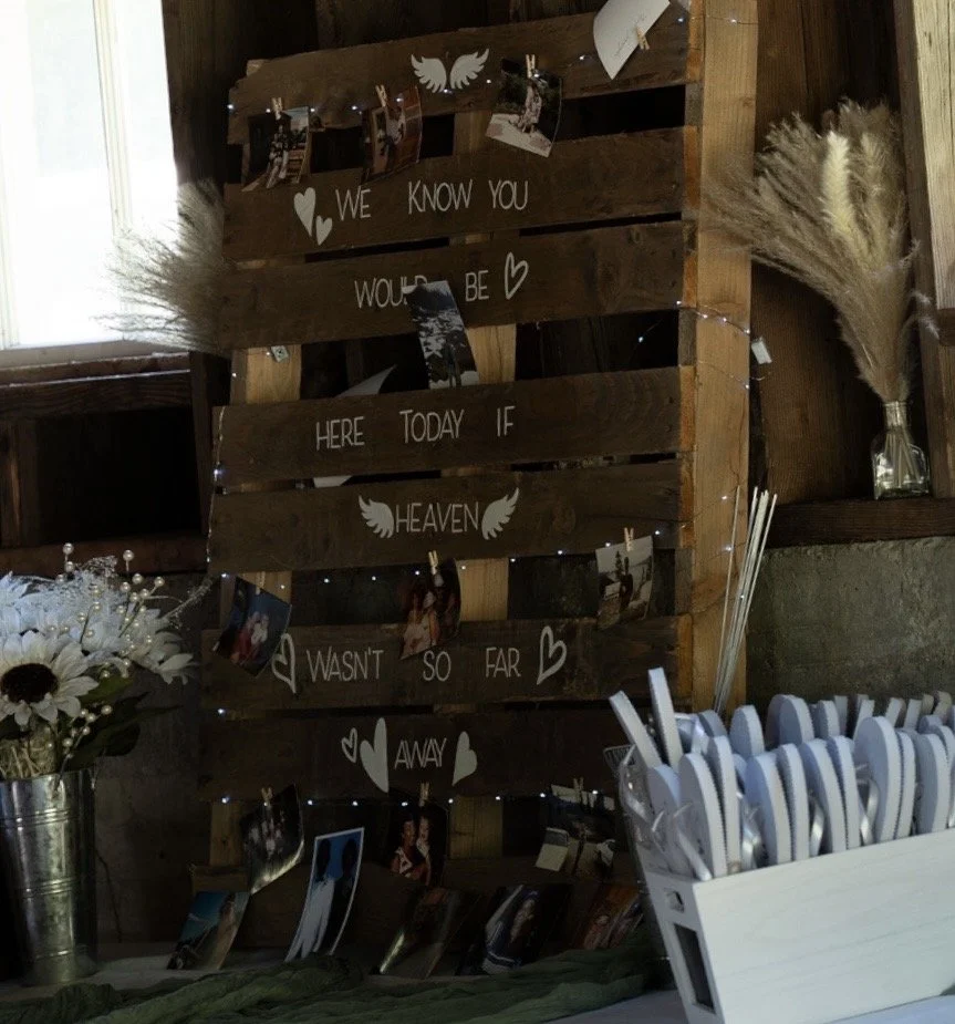 Wooden pallet display with a handwritten message, small photos, and decorative elements for a wedding or memorial event. The message reads: 'We know you would be here today if heaven wasn't so far away.'