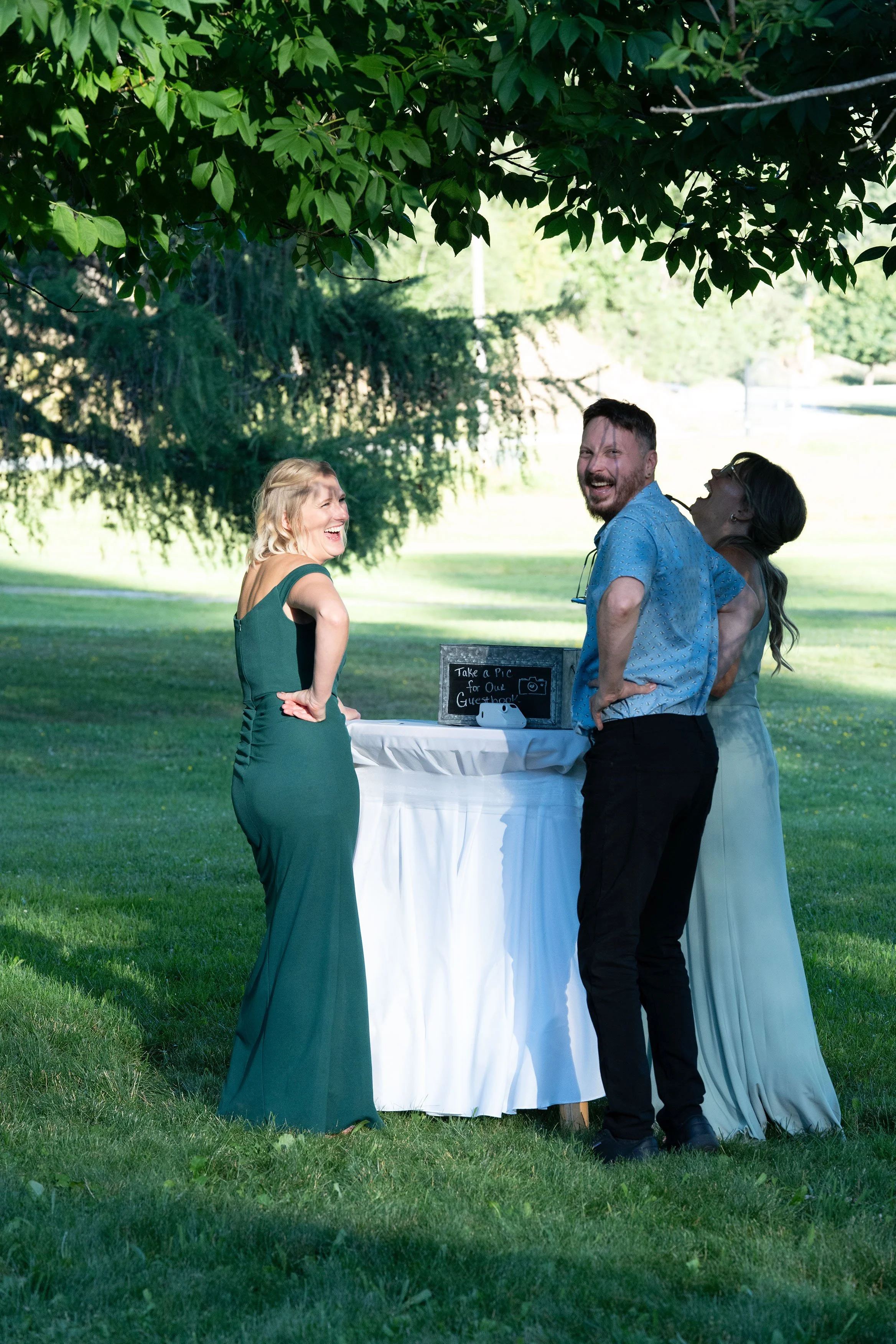 Three people laughing and talking outdoors near a white round table with a sign that reads "Take a Pic for Our Guestbook" under a large tree in a park.